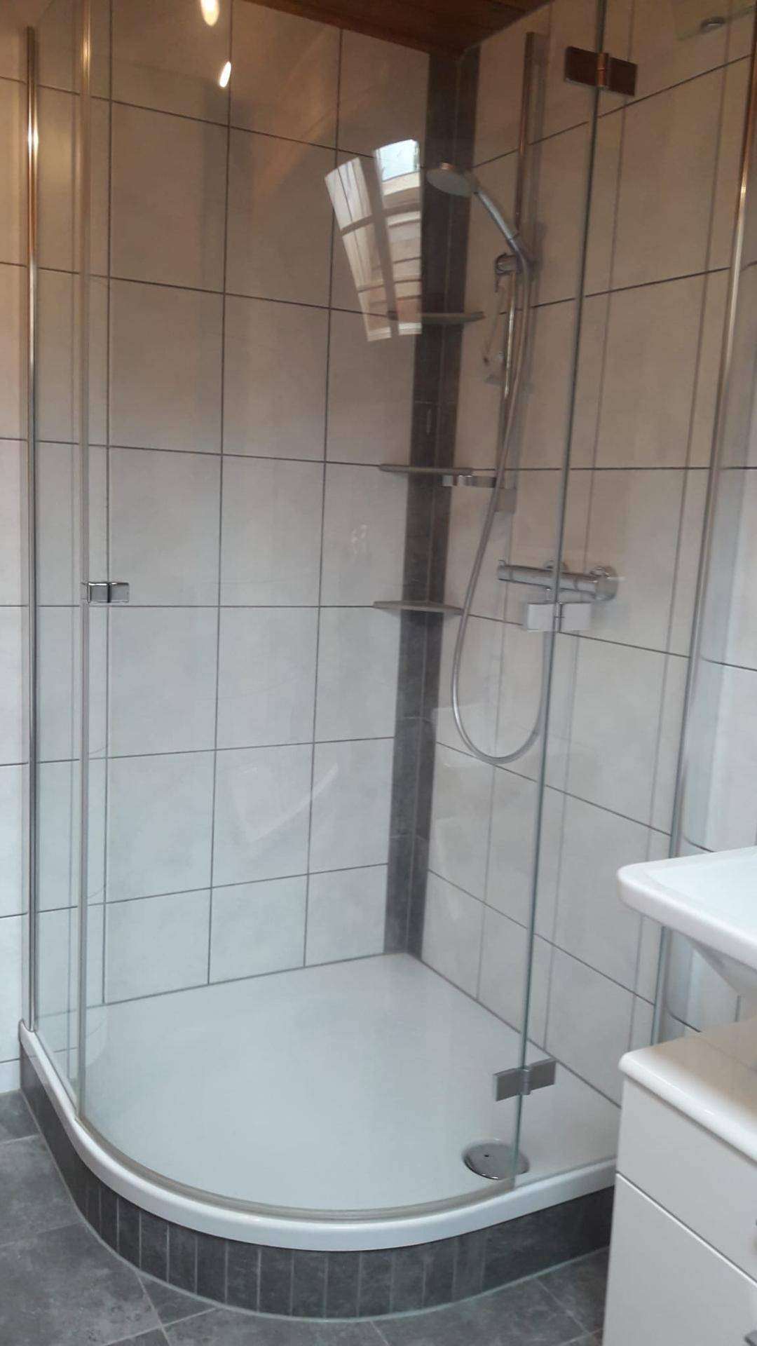 Eckduschkabine mit Glaswänden in einem hell gefliesten Badezimmer mit Holzdecke.