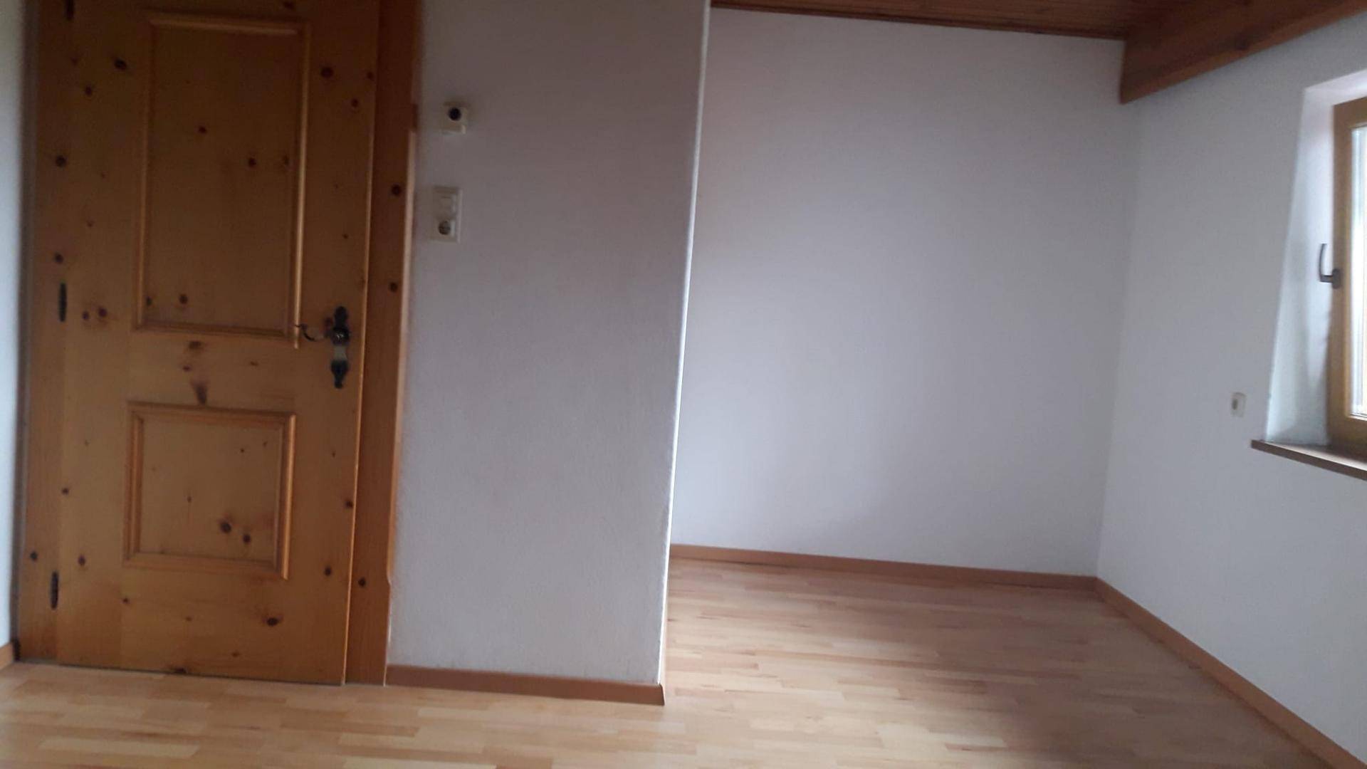 Leeres Zimmer mit hellem Holzboden, weißen Wänden und einem Fenster, das Tageslicht hereinlässt.