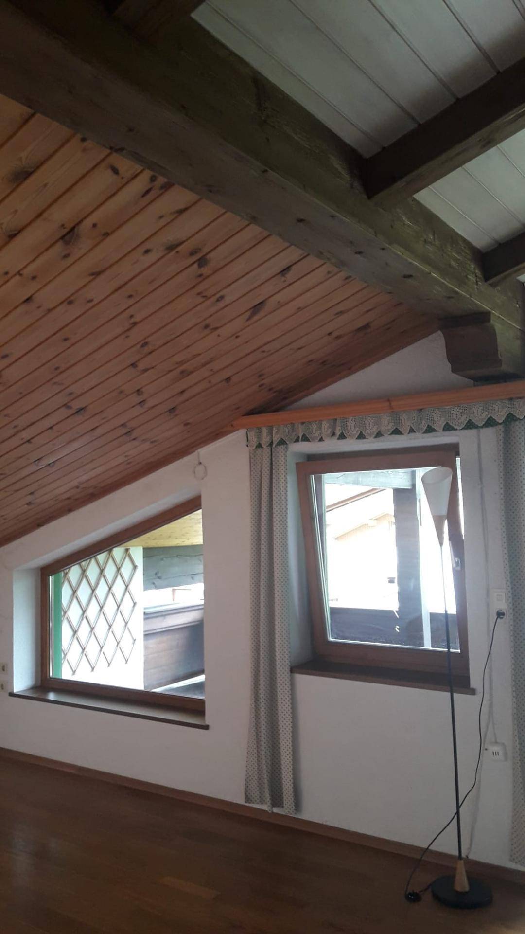 Zimmer mit schräger Holzdecke, Holzboden und zwei Fenstern, die viel natürliches Licht spenden.
