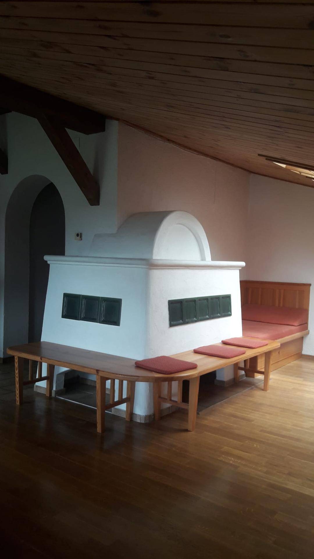 Gemütlicher Wohnbereich mit traditionellem Kachelofen, Holzbänken und sichtbaren Holzbalken an der Decke.