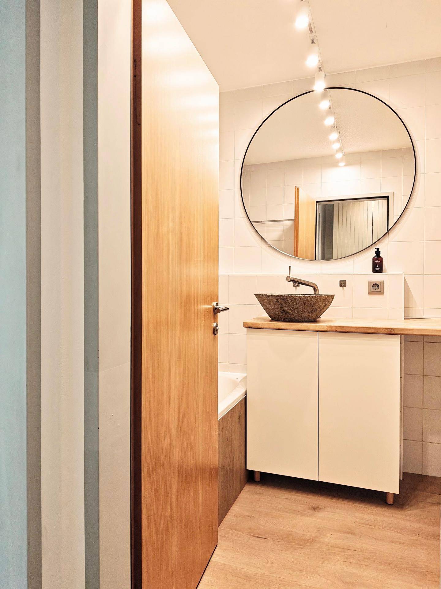 Modernes Badezimmer mit rundem Spiegel, Steinwaschbecken auf Holzplatte und weißen Fliesen.