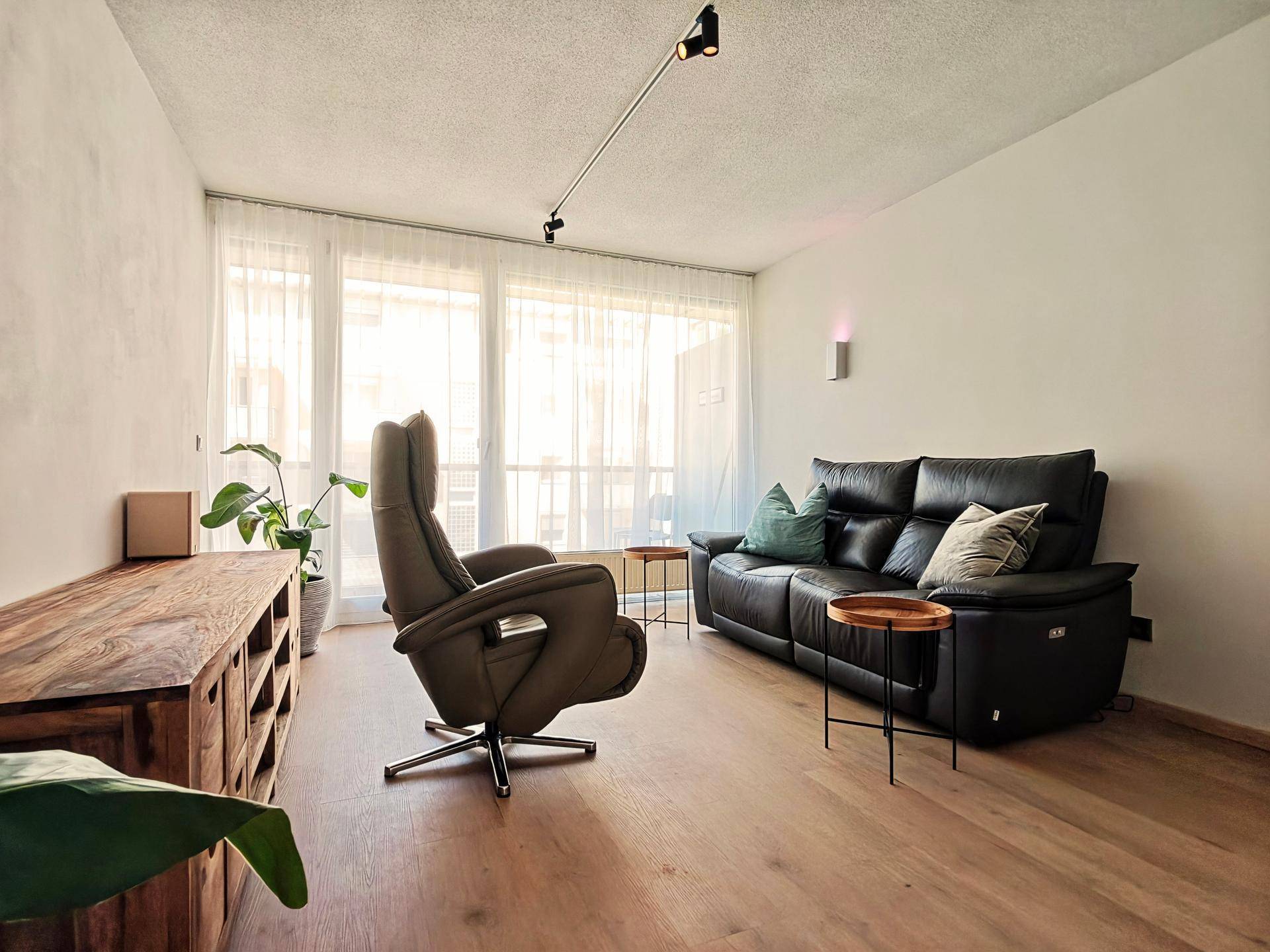 Heller Wohnbereich mit komfortabler Couch, Sessel und einem Holzschrank, ergänzt durch große Fenster.