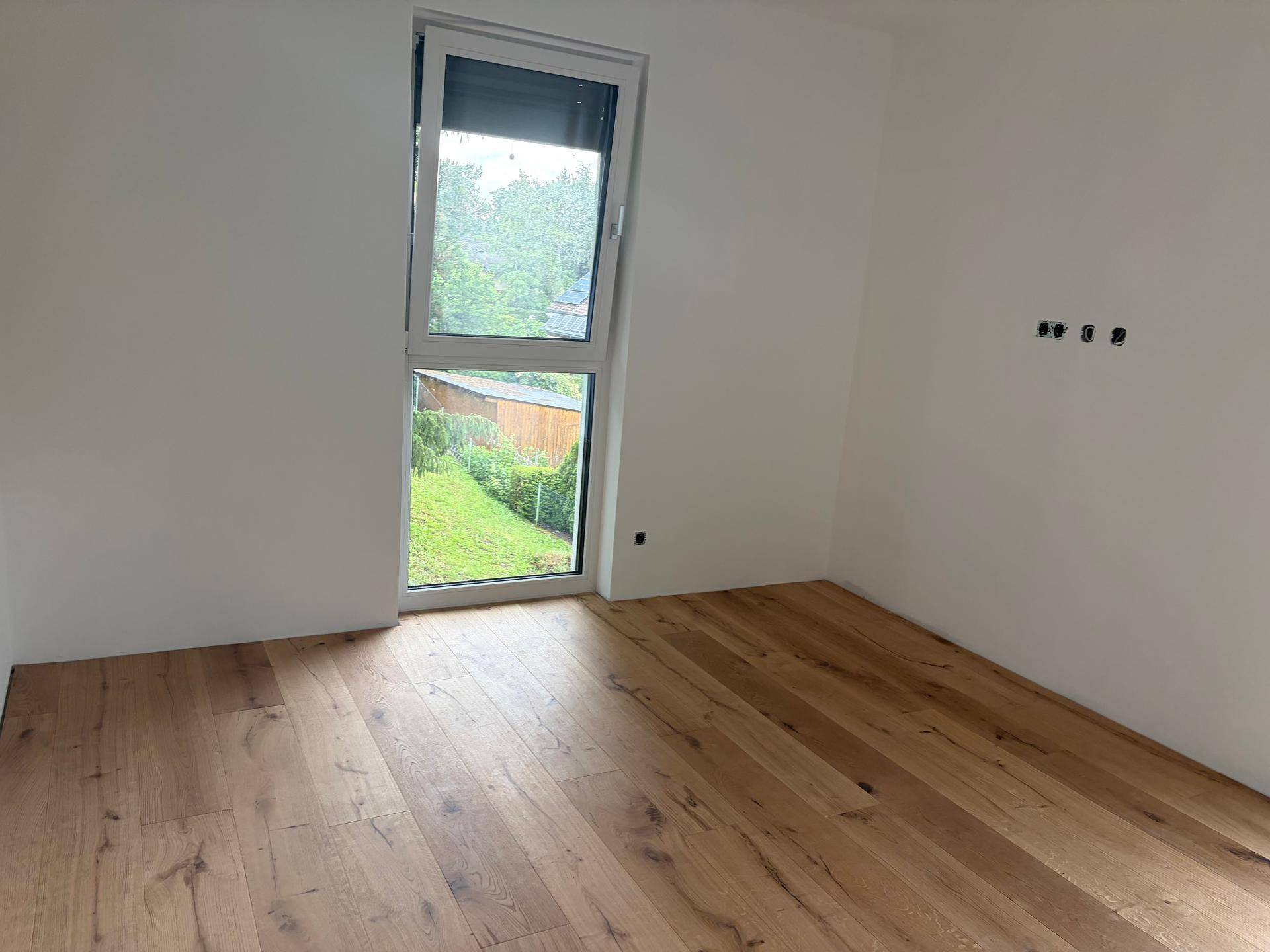 Unmöblierter Raum mit Holzboden und Fenster mit Blick ins Grüne.