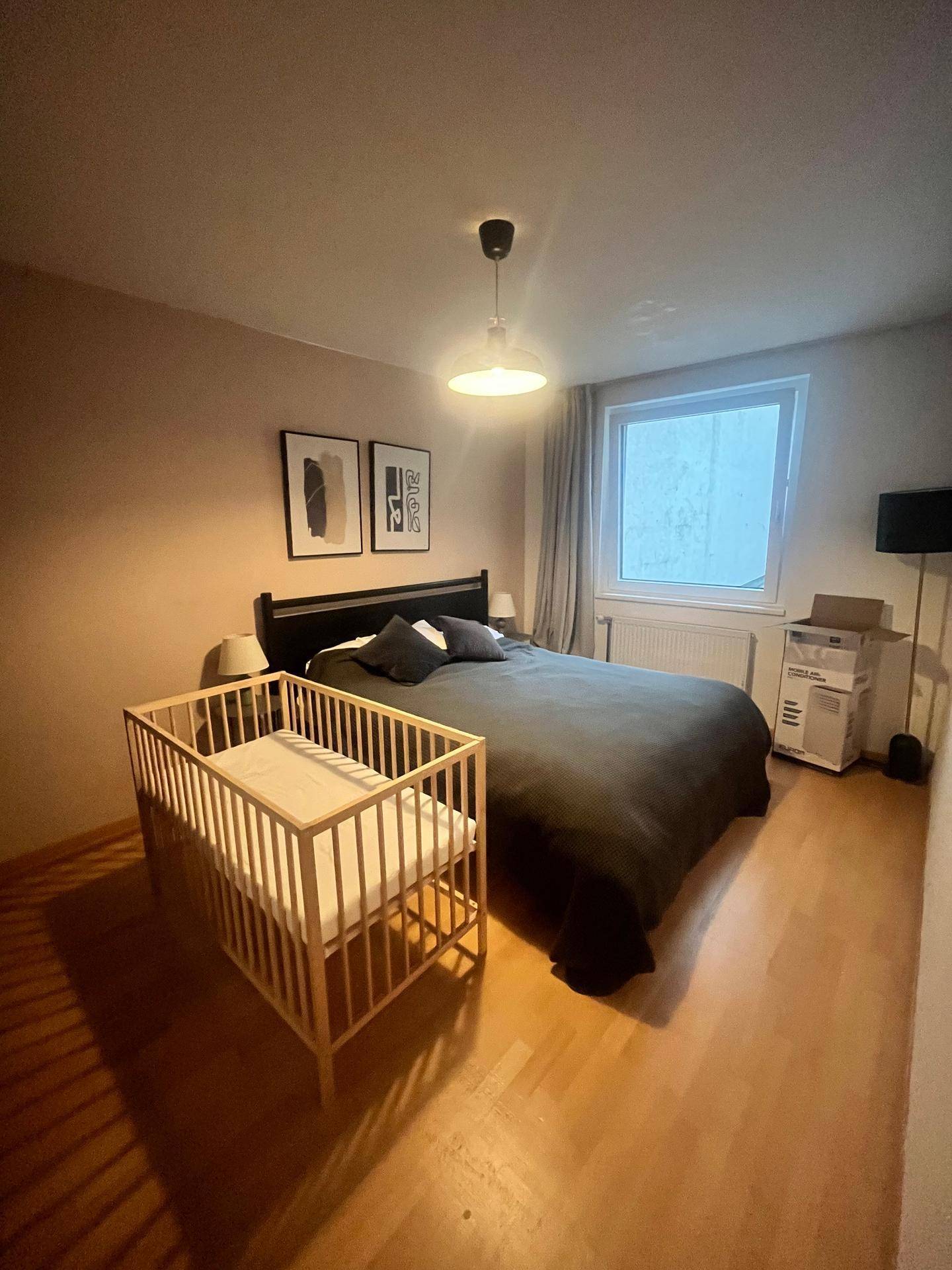 Schlafzimmer mit Holzboden, Doppelbett und Babybett, gut beleuchtet durch ein großes Fenster.