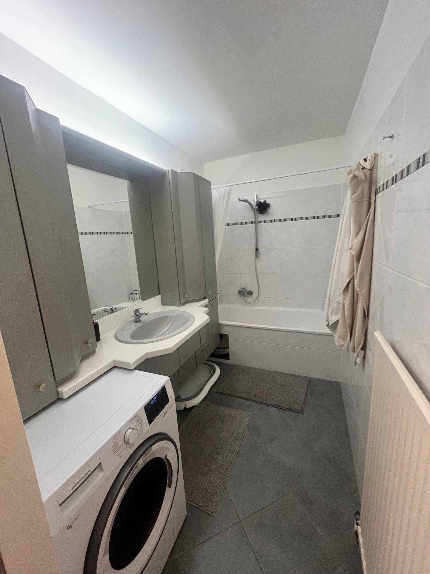 Badezimmer mit Badewanne, Waschbecken, Waschmaschine und grauen Fliesen.