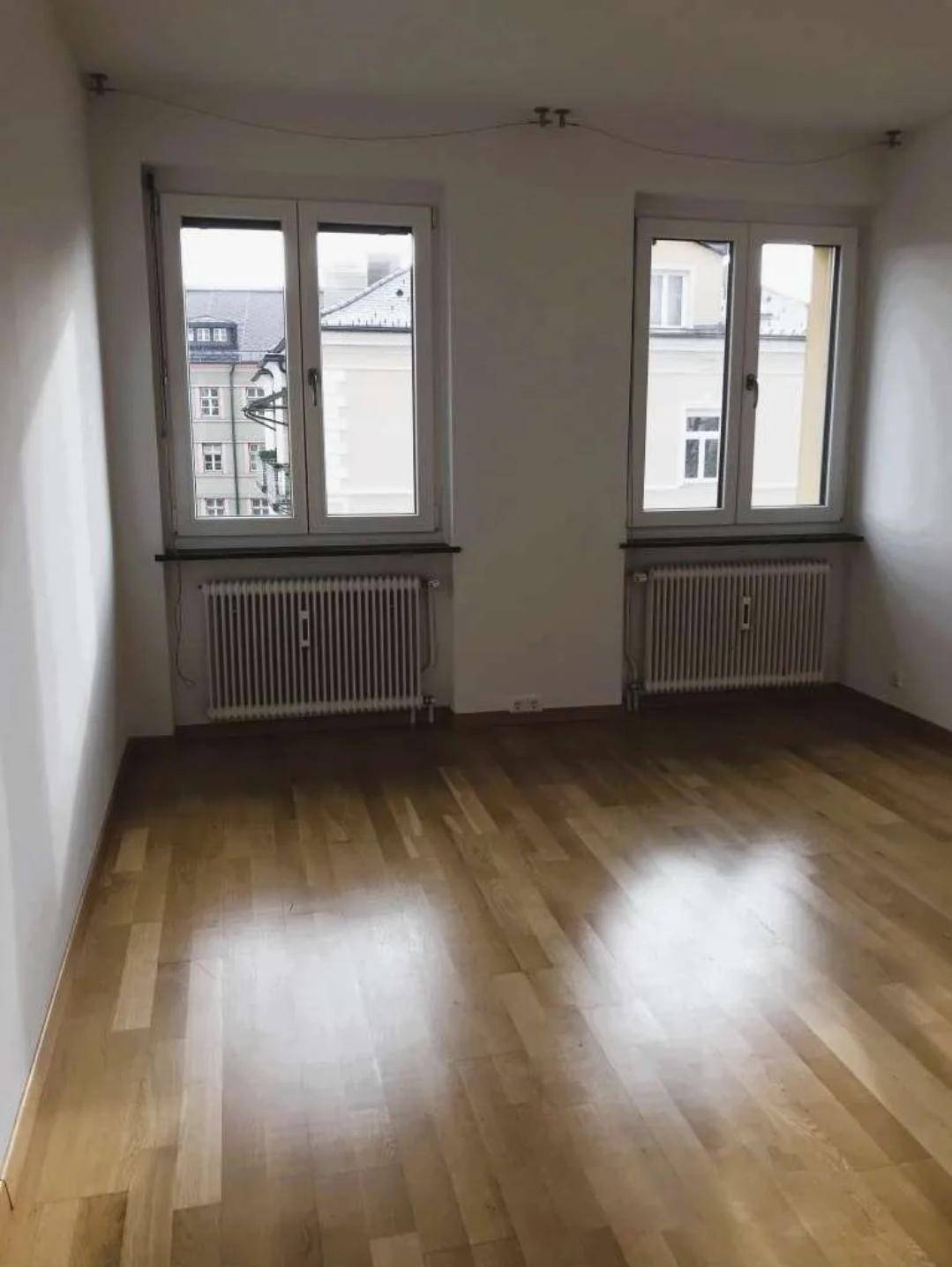 Geräumiges Zimmer mit Holzboden und zwei Fenstern, das viel Tageslicht hereinlässt.