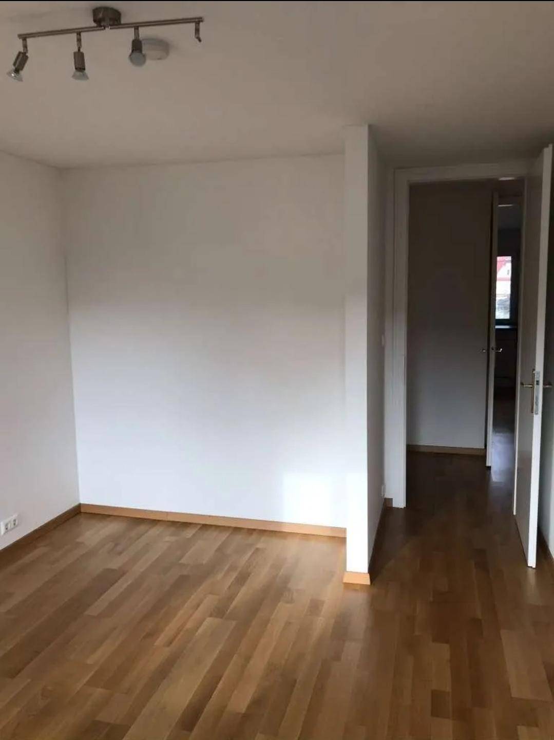 Helles, leeres Zimmer mit Holzboden und Zugang zu einem Flur, bereit zur individuellen Gestaltung.