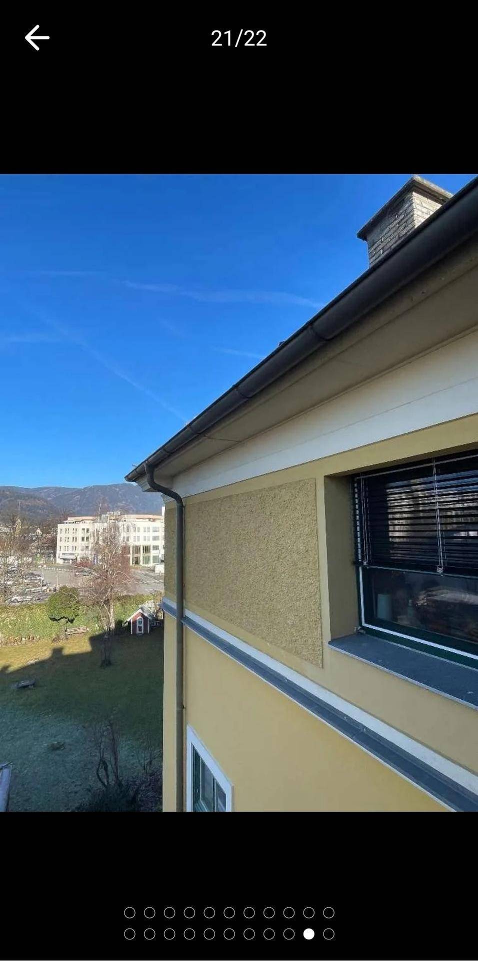 Gebäudeecke mit Fenster und Jalousien, bietet einen Blick auf die Umgebung und entfernte Berge.