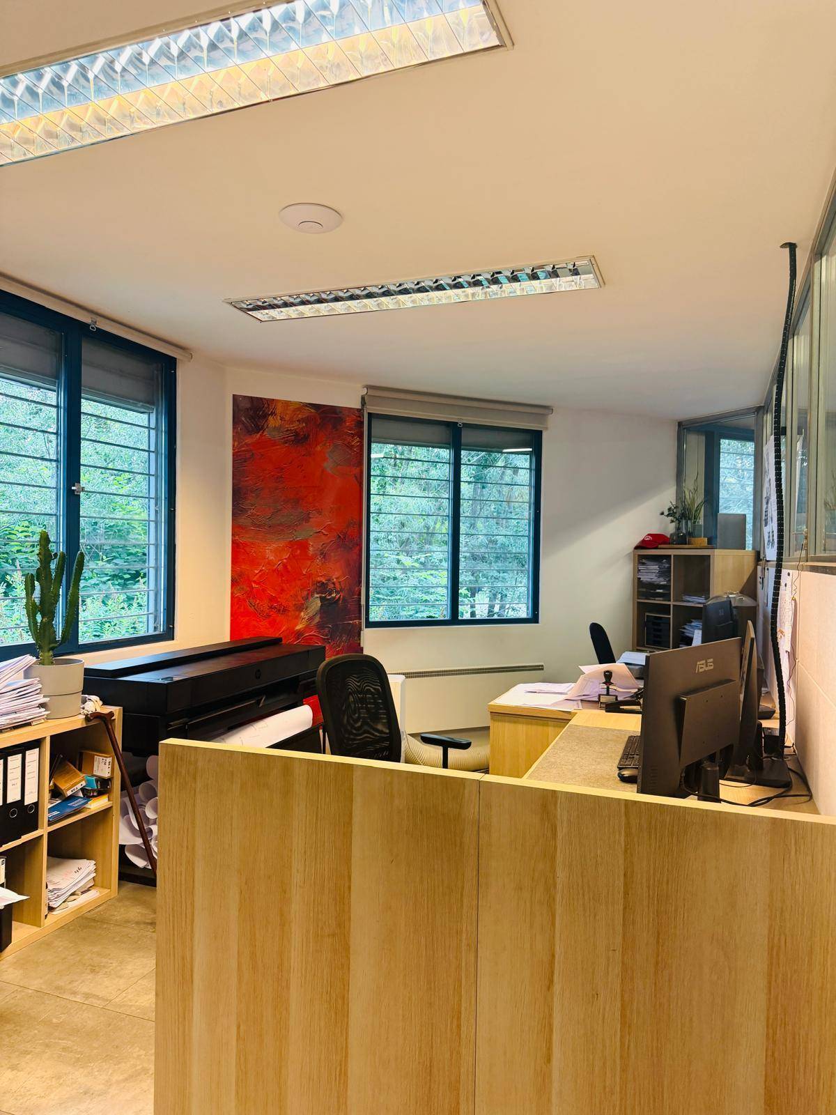 Modernes Büro mit Holzmöbeln, großen Fenstern und einem roten Kunstwerk an der Wand, sehr hell.