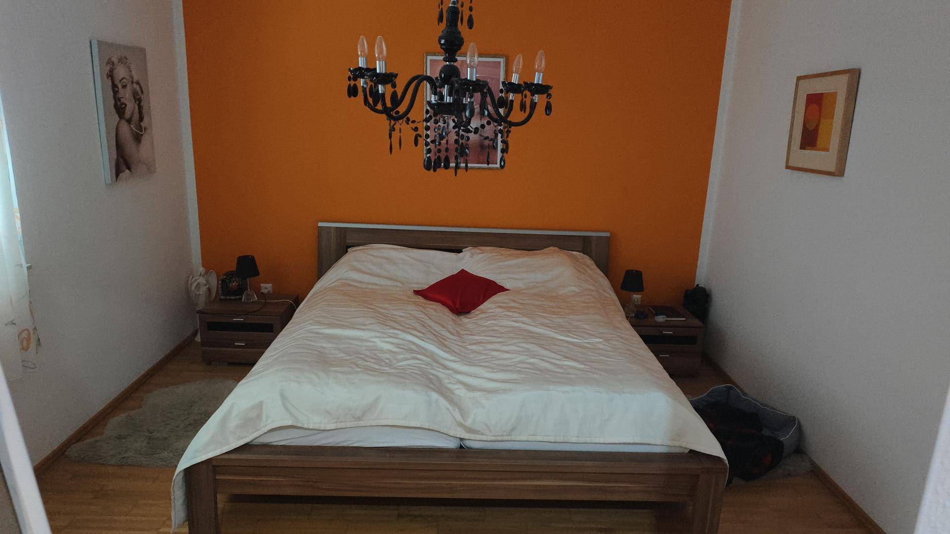 Schlafzimmer mit orangefarbener Akzentwand, Holzbett und einem dunklen Kronleuchter.