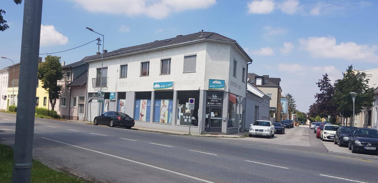 Gebäudeansicht von der Straßenecke mit Geschäftsflächen im Erdgeschoss und Parkplätzen.