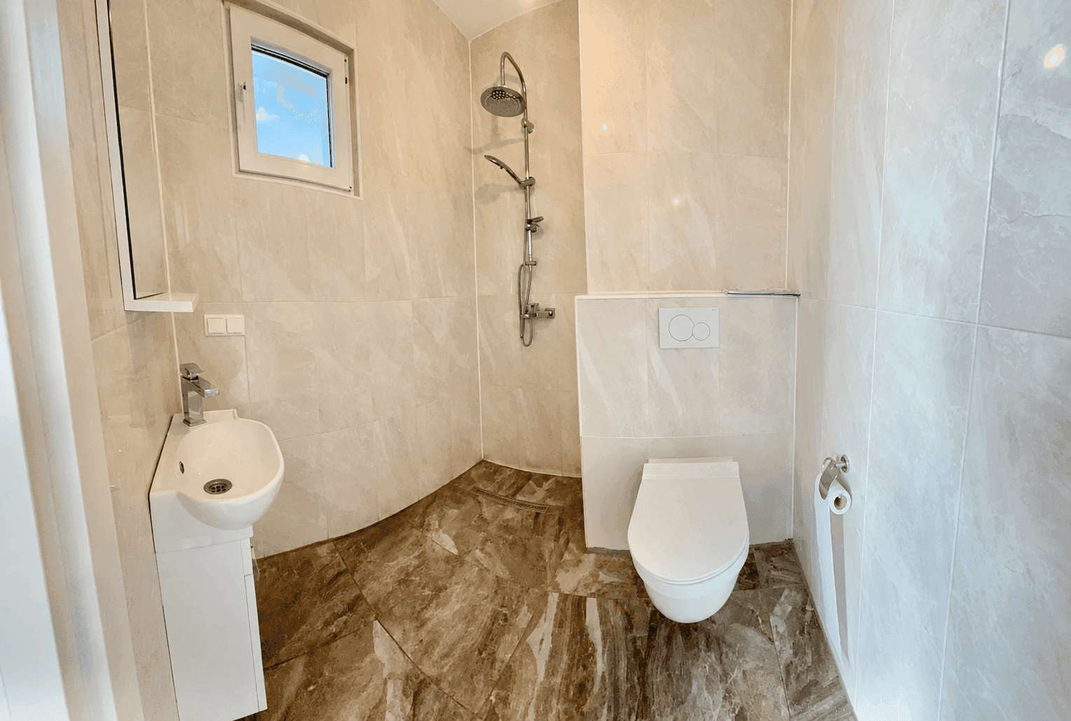 Modernes Badezimmer mit beigefarbenen Fliesen, Dusche, wandhängendem WC und kleinem Waschbecken.