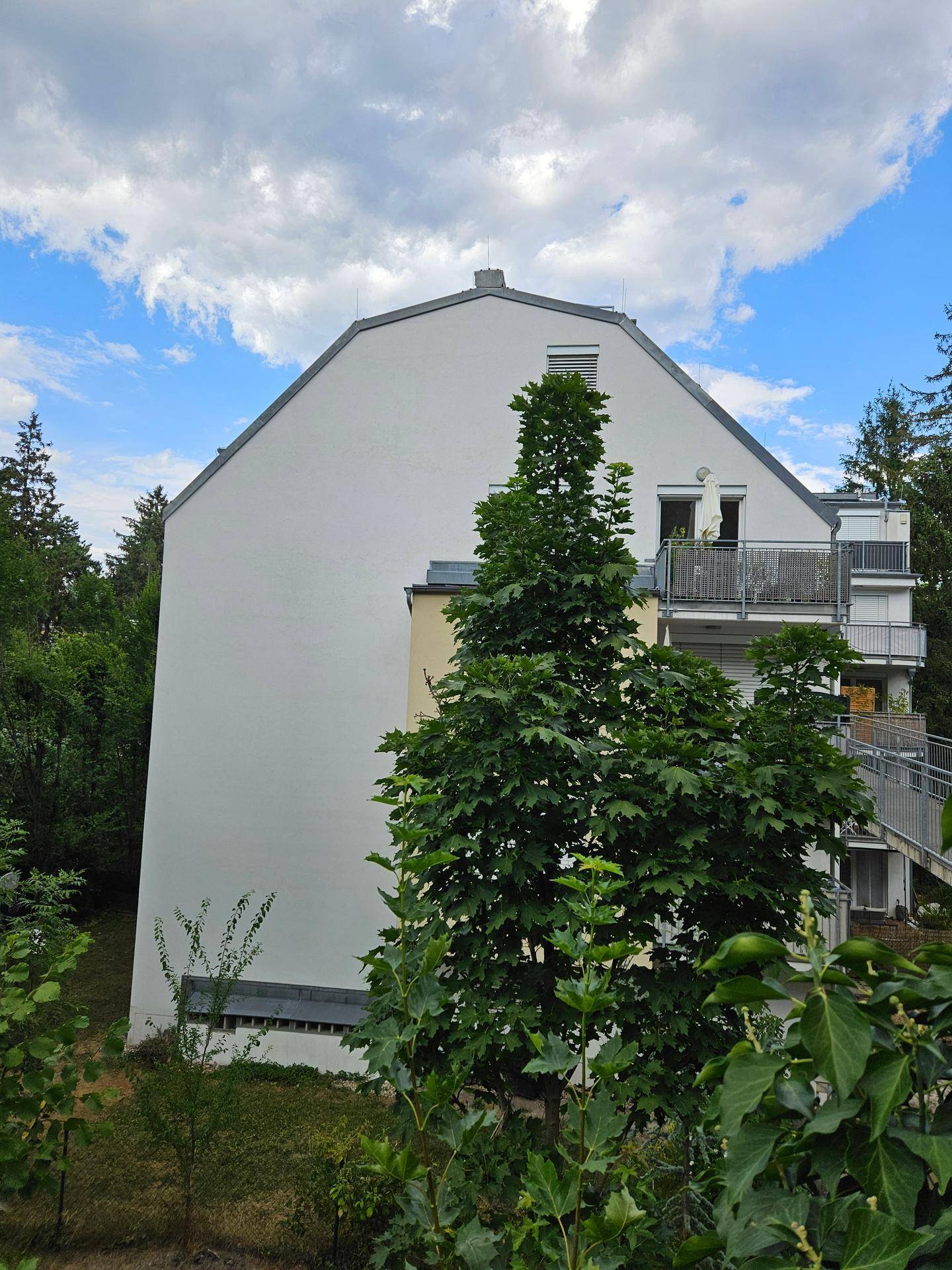 Weißes Mehrfamilienhaus mit Satteldach und mehreren Balkonen, umgeben von Bäumen.