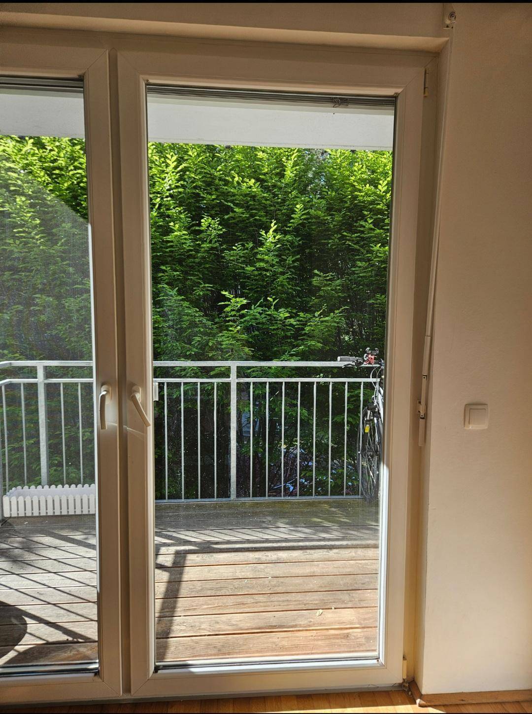 Blick durch die Balkontür auf die Holzterrasse und das umgebende grüne Blätterdach.