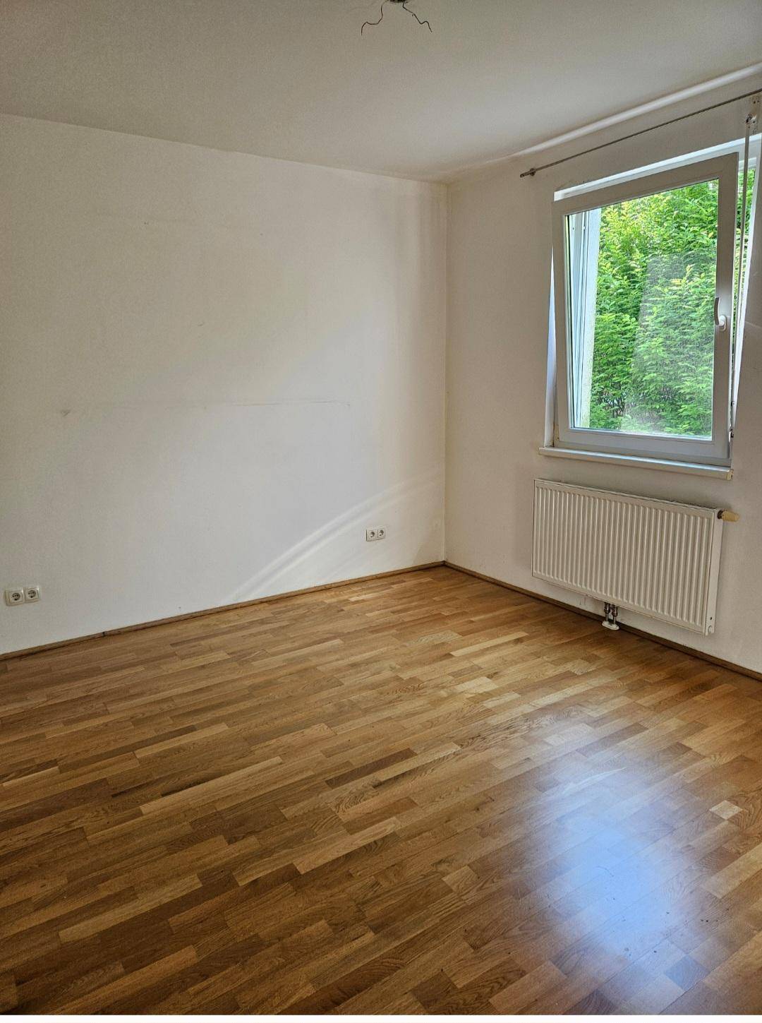 Geräumiges Zimmer mit Parkettboden, weißen Wänden und einem Fenster, das Tageslicht hereinlässt.