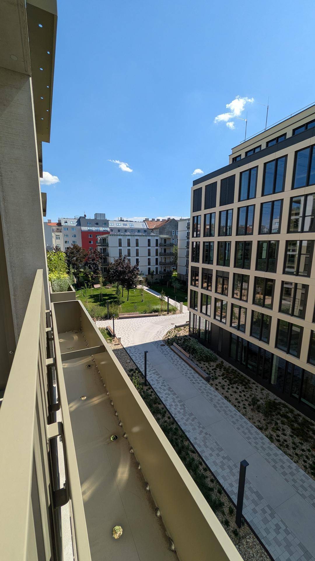 Blick von einem Balkon auf einen begrünten Innenhof und moderne Wohngebäude unter blauem Himmel.