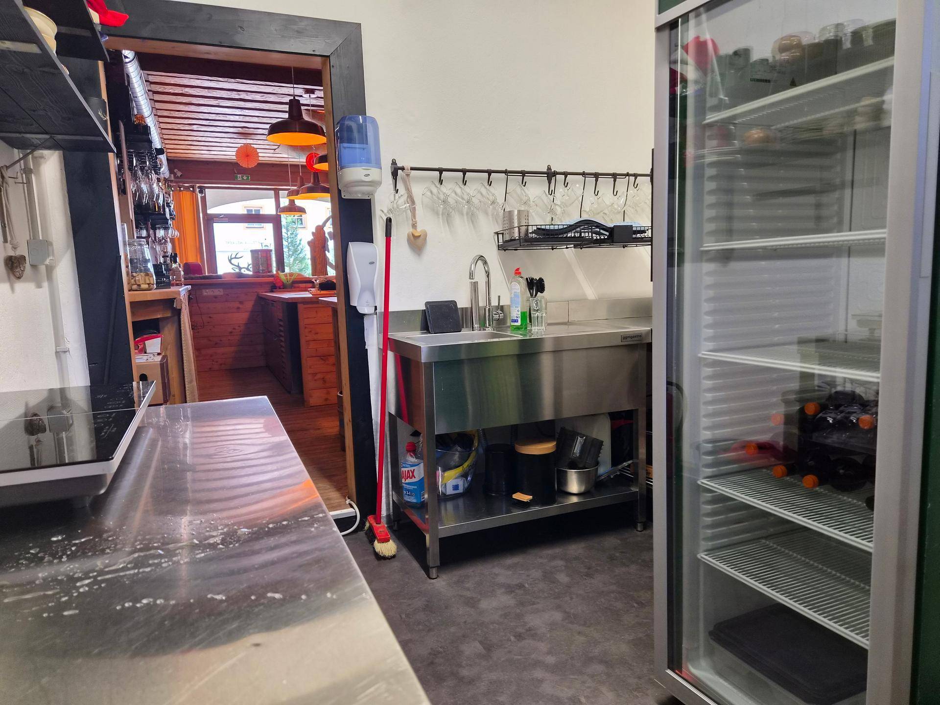 Gewerbliche Küche mit Edelstahlspüle, großem Kühlschrank und Blick in den Barbereich.