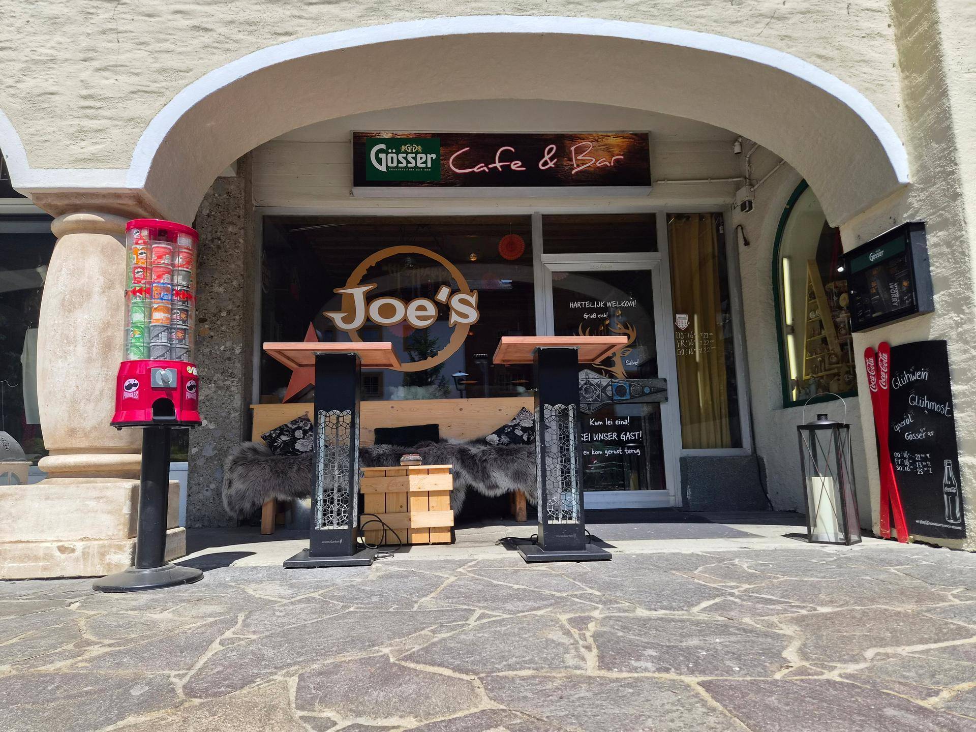 Eingangsbereich eines Cafés mit Außentischen und einem Werbeschild auf dem Bürgersteig.
