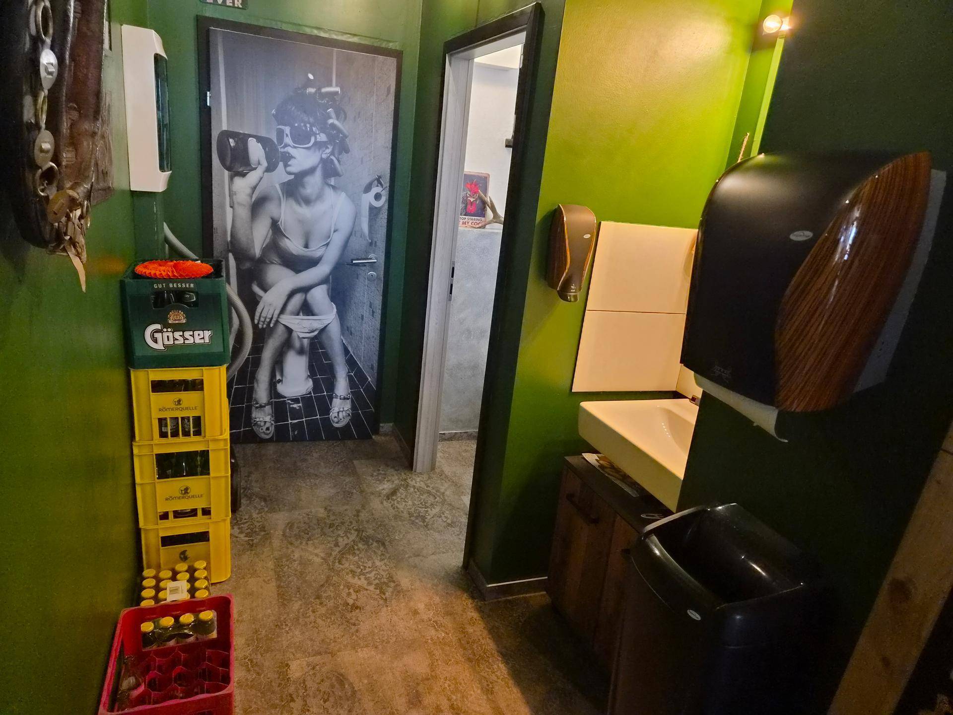 Flur zu den Toiletten mit grünen Wänden, Wickeltisch und einer auffälligen Türdekoration.