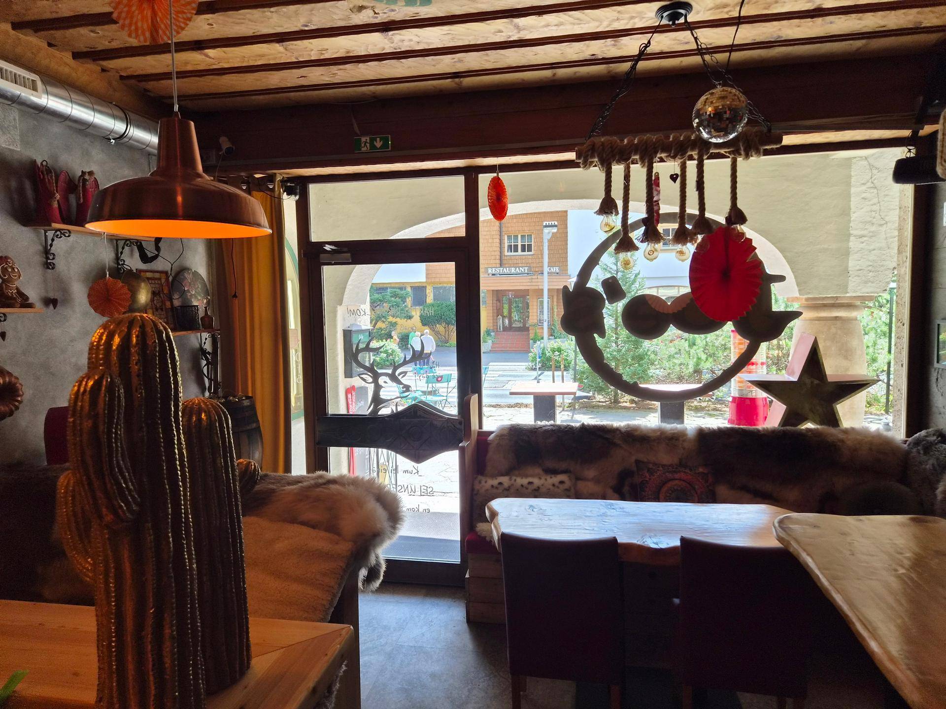 Innenansicht des Cafés mit Blick auf den Eingang, Sitzgelegenheiten und die Außenterrasse durch große Fenster.