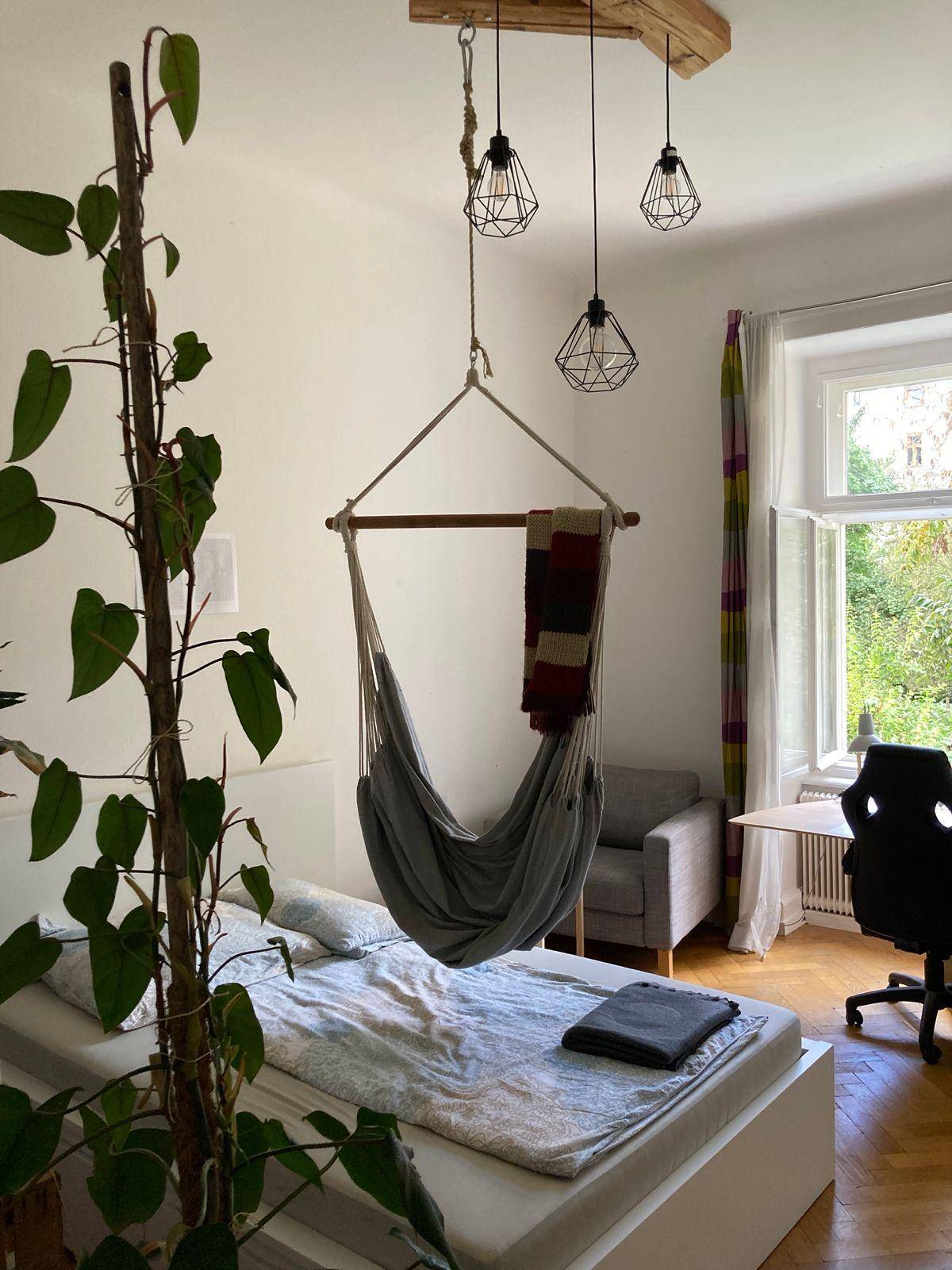 Helles Schlafzimmer mit Doppelbett, Hängesessel und Blick ins Grüne durch das Fenster.
