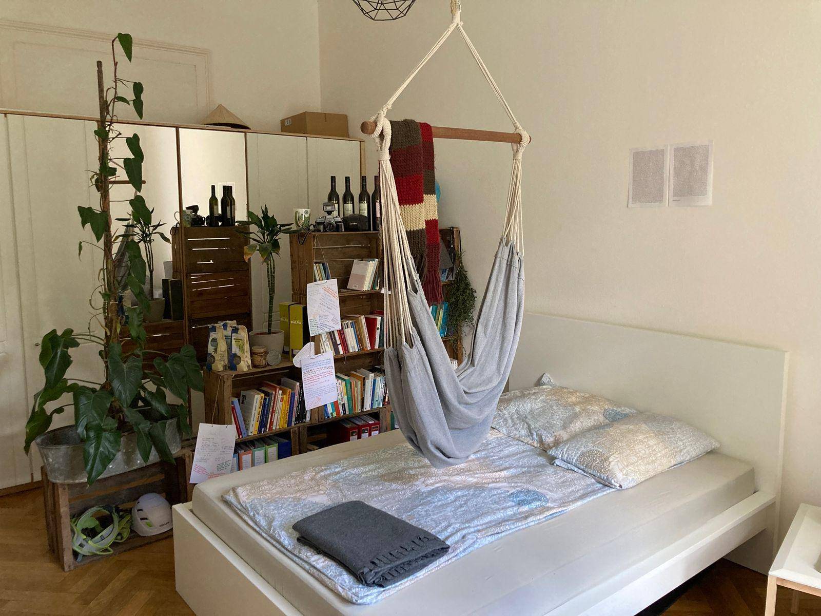Helles Schlafzimmer mit Hängesessel, großem Bett und kreativem Bücherregal aus Holzkisten.