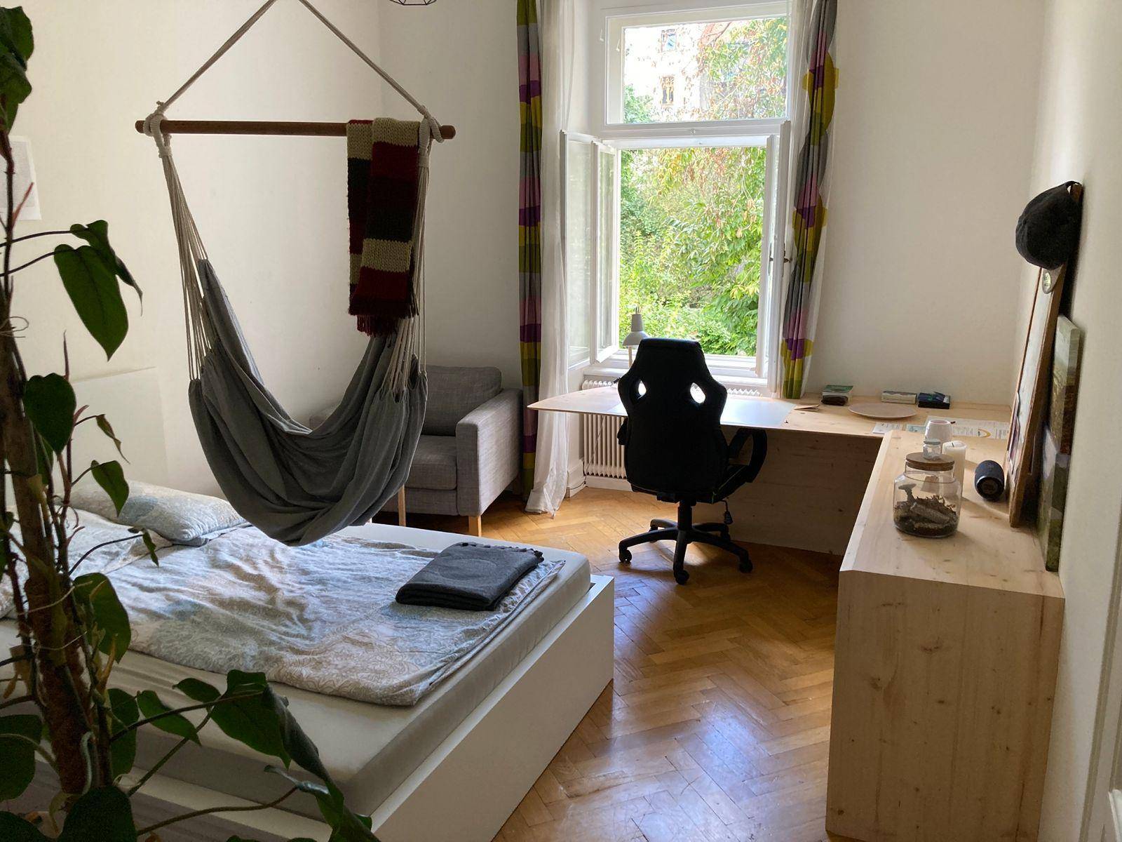 Geräumiges Schlafzimmer mit Bett, Hängesessel und Arbeitsbereich am Fenster mit Grünblick.