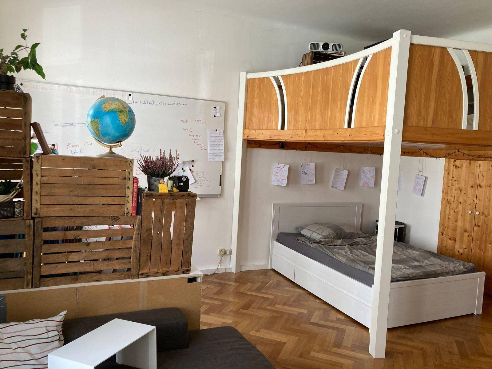 Schlafzimmer mit Hochbettkonstruktion, darunter ein Bett und ein Bücherregal aus Holzkisten.
