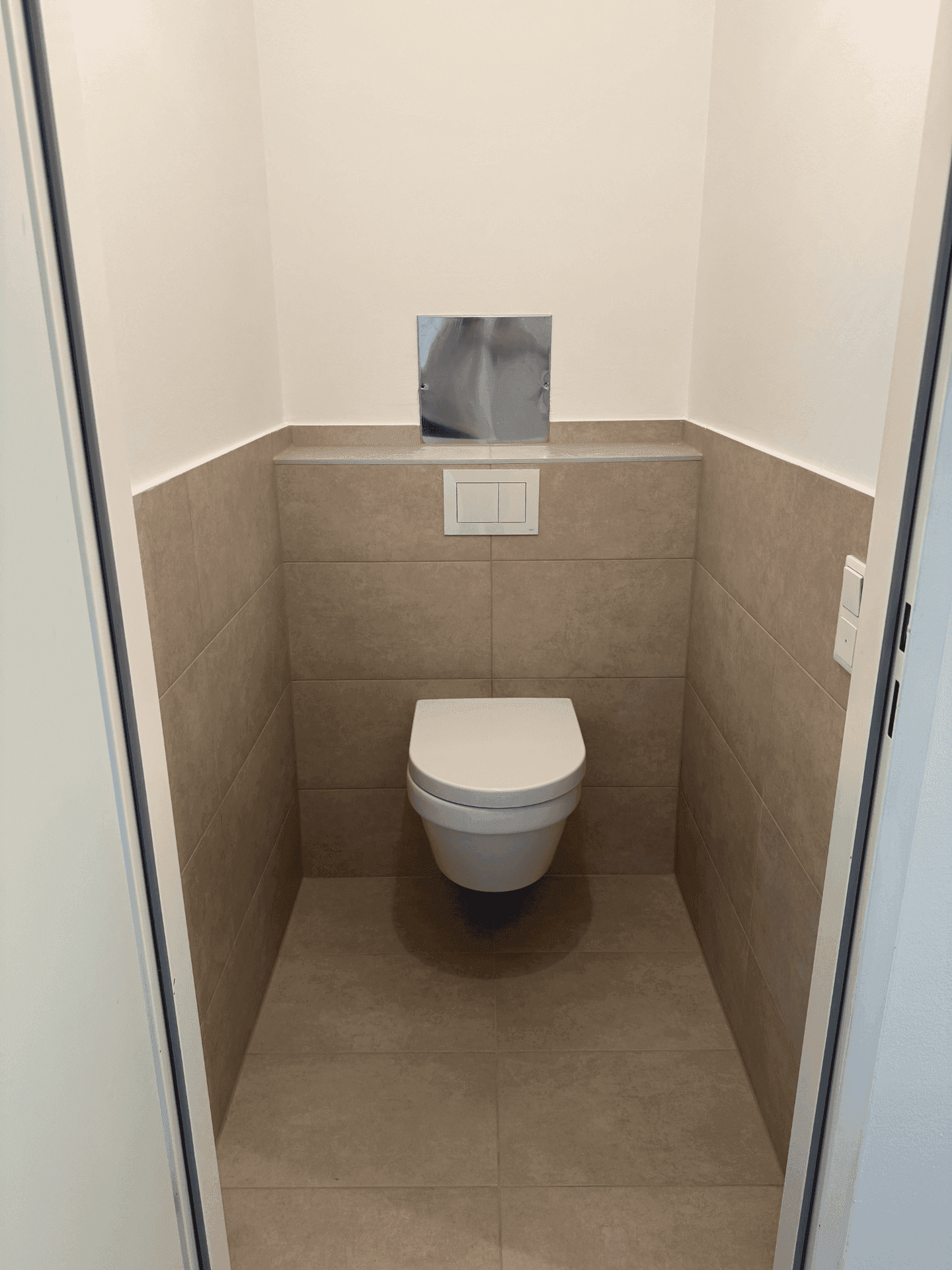 Separates WC mit wandhängender Toilette und neutralen Fliesen an Wänden und Boden.