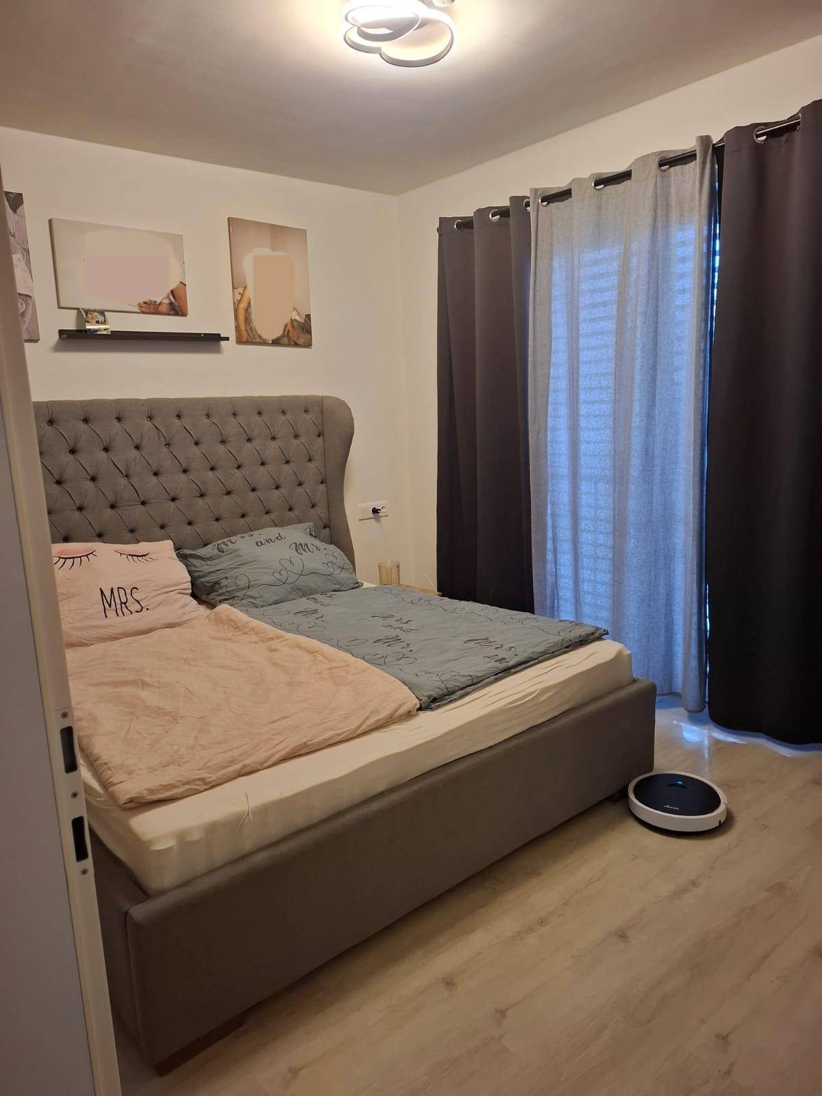 Gemütliches Schlafzimmer mit großem Polsterbett, dunklen Vorhängen und hellem Laminatboden.