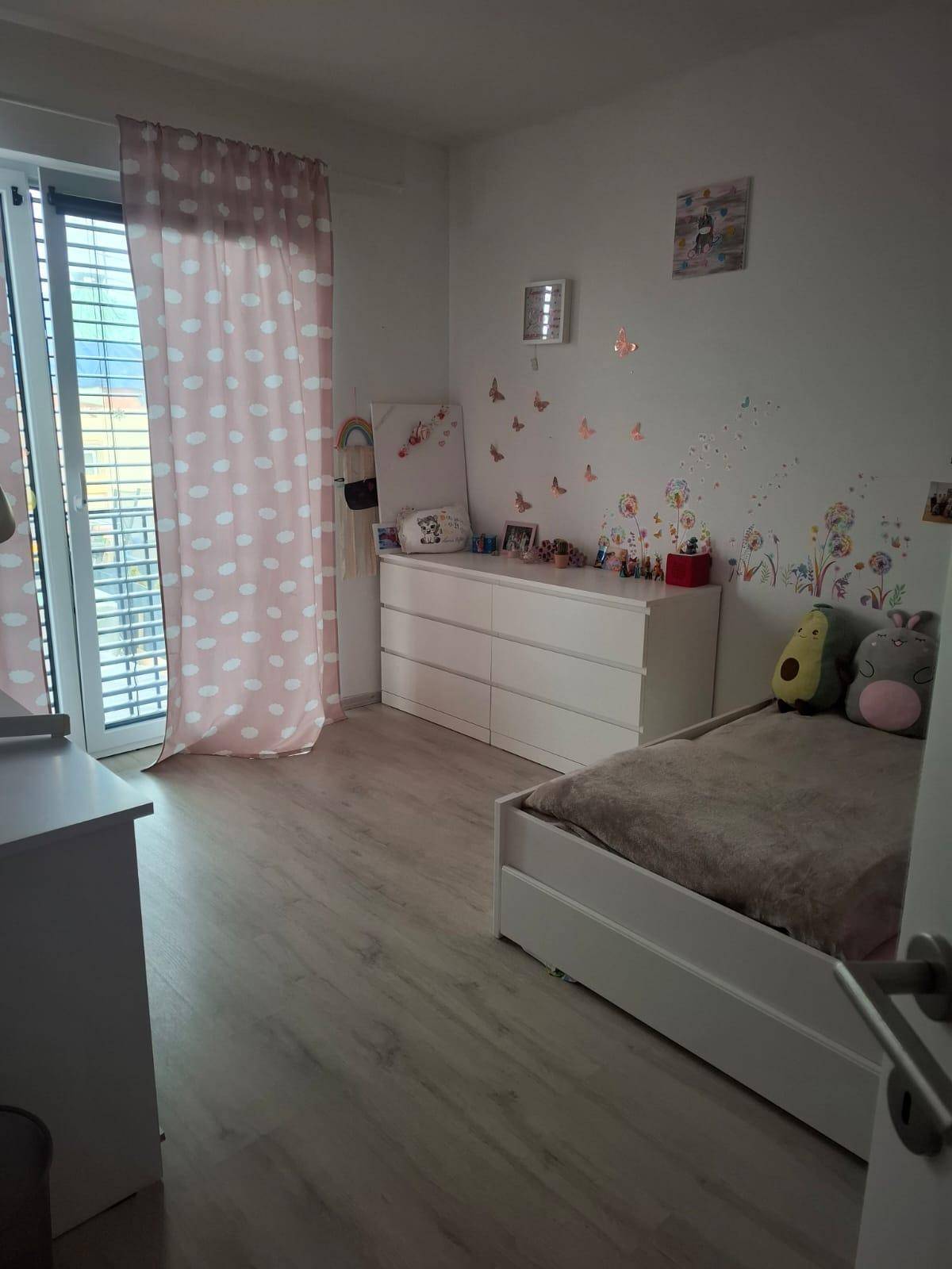 Kinderzimmer mit hellem Laminatboden, Kommode und Zugang zu einem französischen Balkon.