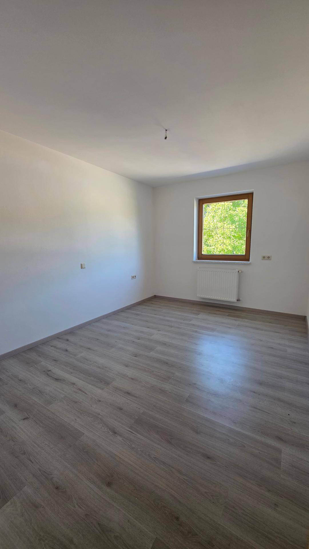Leeres Zimmer mit Laminatboden, weißen Wänden und einem Fenster mit Blick auf Bäume.