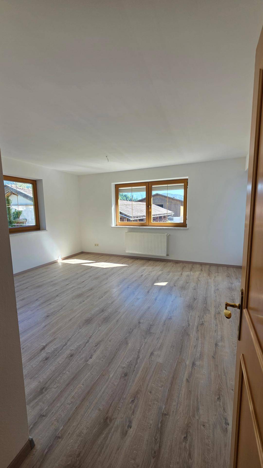 Helles, leeres Zimmer mit Laminatboden, weißen Wänden und großen Fenstern.