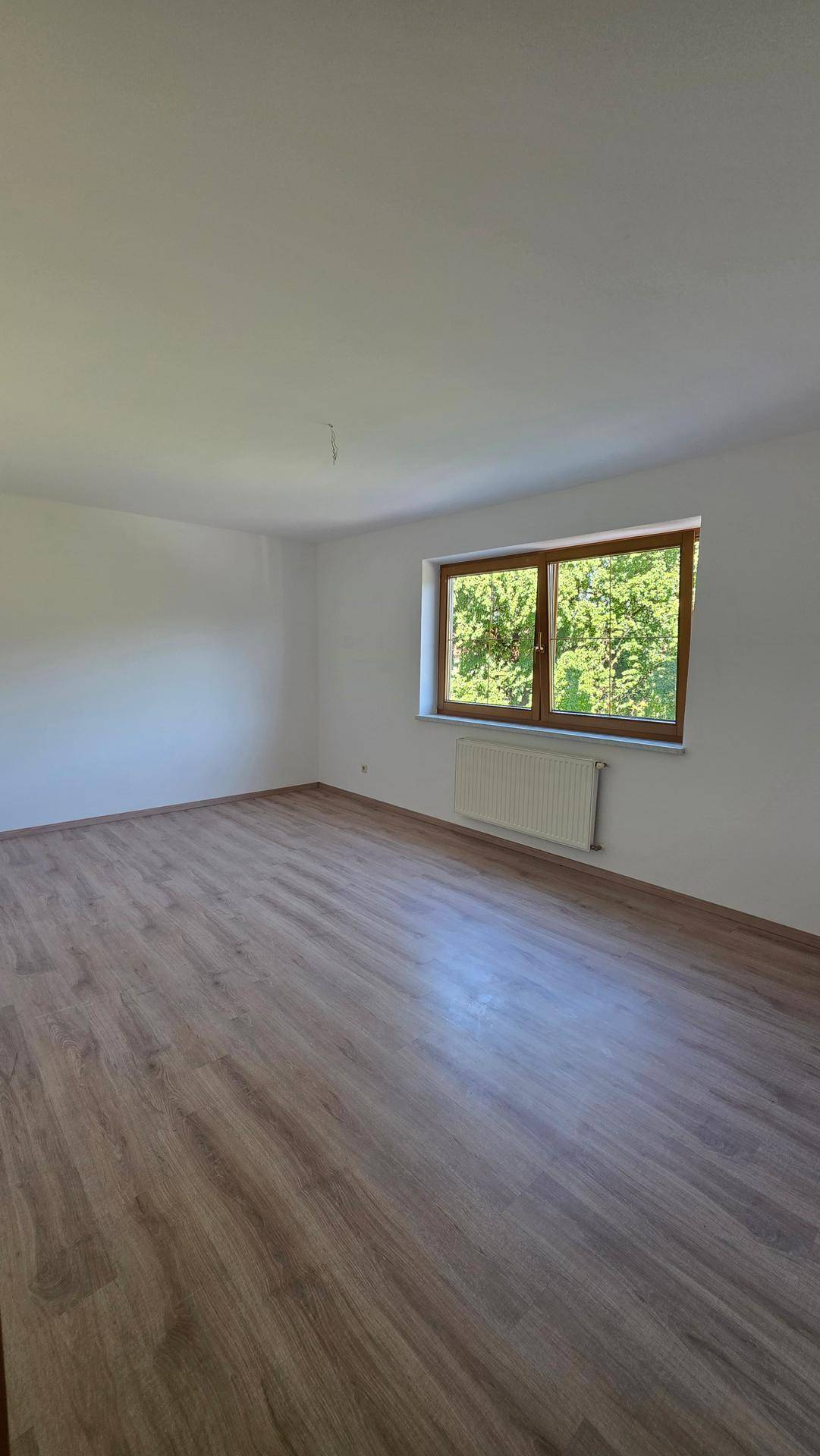 Leeres Zimmer mit Laminatboden, weißen Wänden und einem Fenster mit Blick ins Grüne.