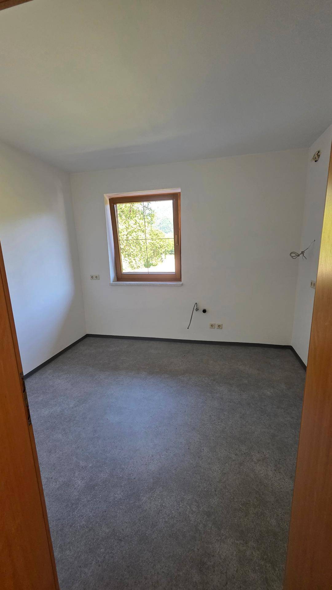 Unmöbliertes Zimmer mit grauem Bodenbelag, weißen Wänden und einem Fenster mit grüner Aussicht.