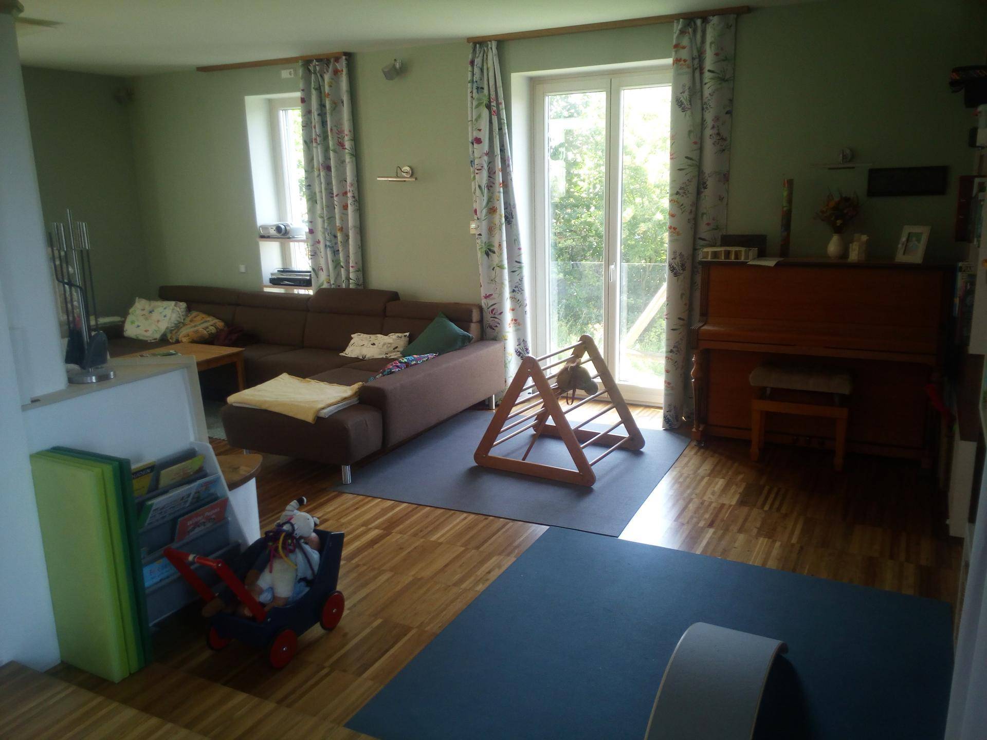 Helles Wohnzimmer mit Sofa, Klavier und großen Fenstern mit Zugang zum Außenbereich.