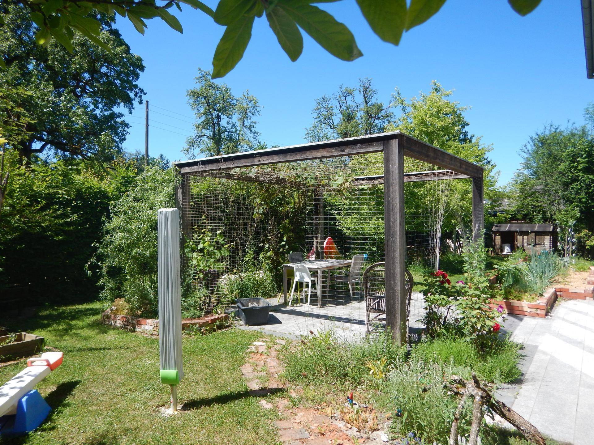 Idyllischer Gartenbereich mit einer Holzpergola, Esstisch und üppiger Vegetation.
