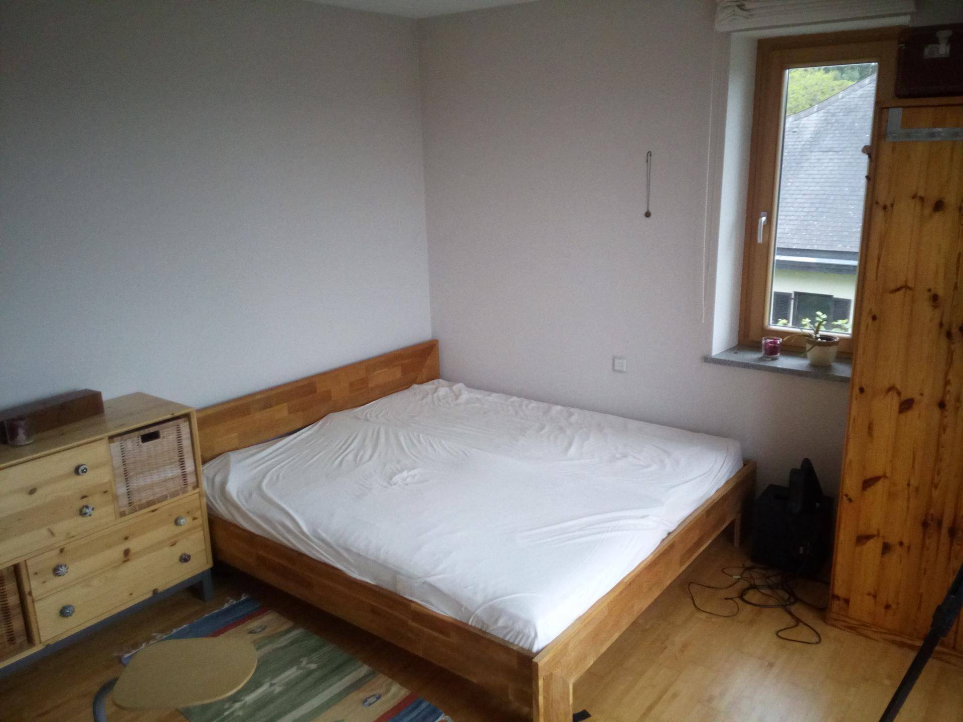 Gemütliches Schlafzimmer mit Holzbett, Kommode und Kleiderschrank, gut beleuchtet durch ein Fenster.