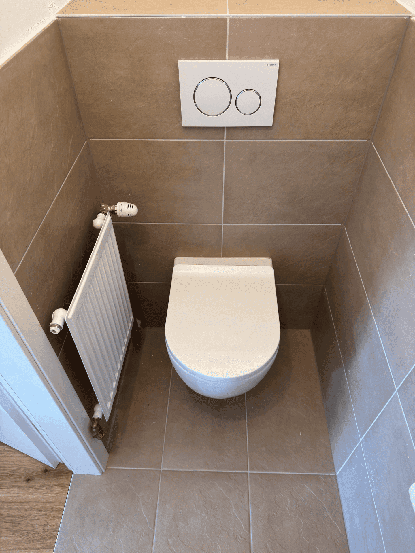 Kleines WC mit braunen Fliesen an Wänden und Boden, ausgestattet mit einer Wandtoilette und Heizkörper.