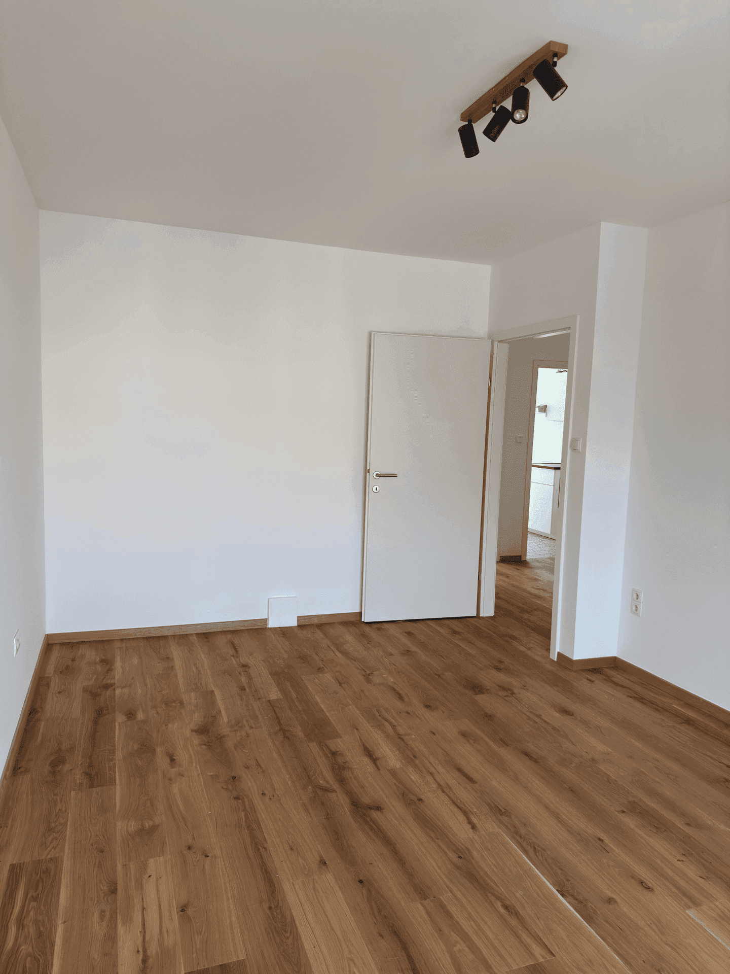 Heller, leerer Raum mit Holzboden und weiß gestrichenen Wänden, mit offener Tür zu einem weiteren Zimmer.