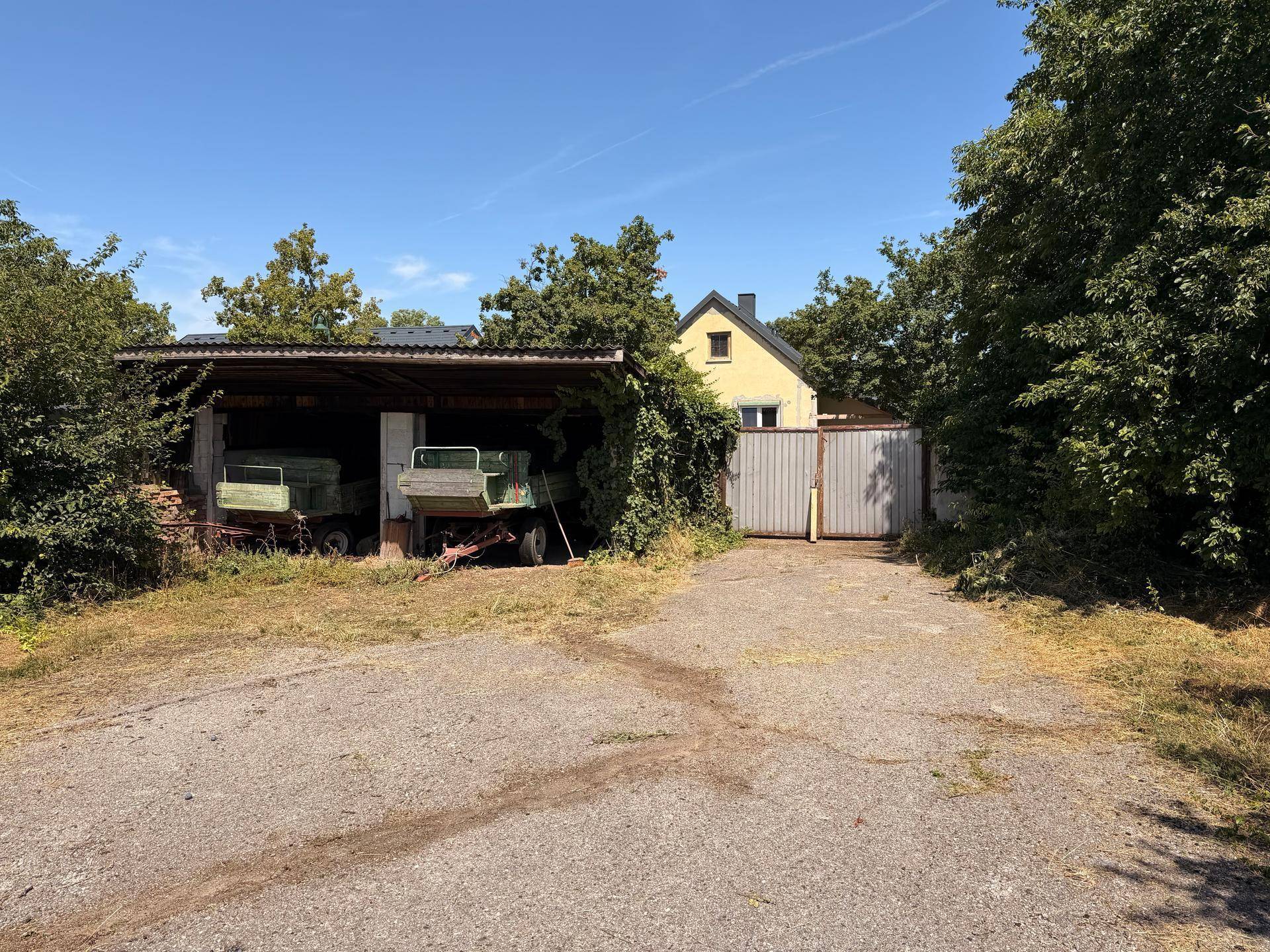 Weitläufige Zufahrt mit Bäumen, einem überdachten Unterstand und einem gelben Haus.