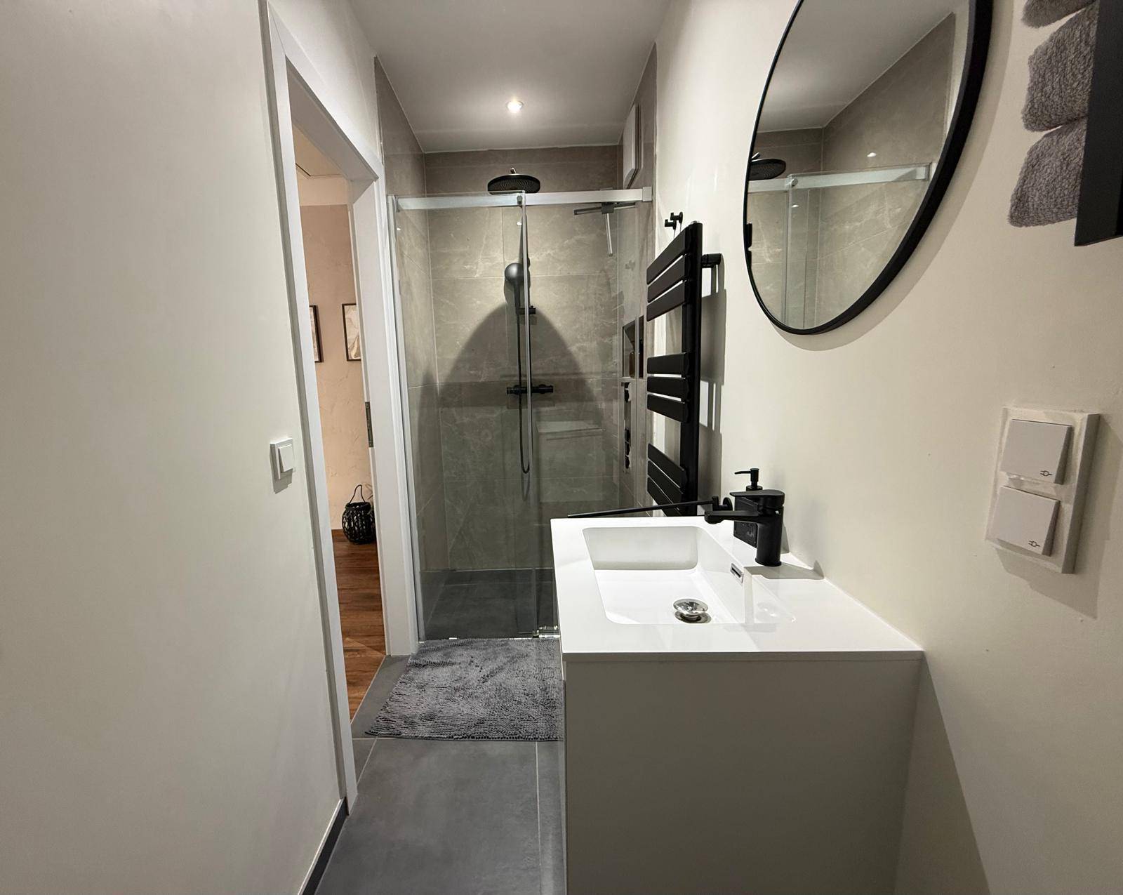 Modernes Badezimmer mit begehbarer Dusche, schwarzem Heizkörper und rundem Spiegel.