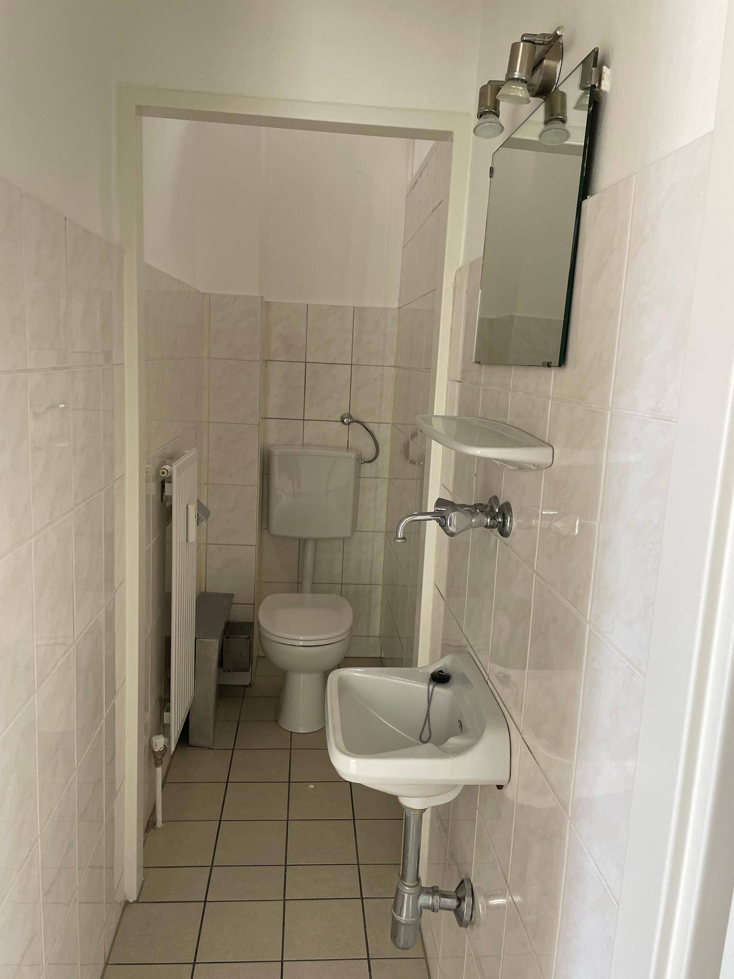 Kleiner Toilettenraum mit weißen Fliesen, Waschbecken und Spiegel, in gutem und sauberem Zustand.