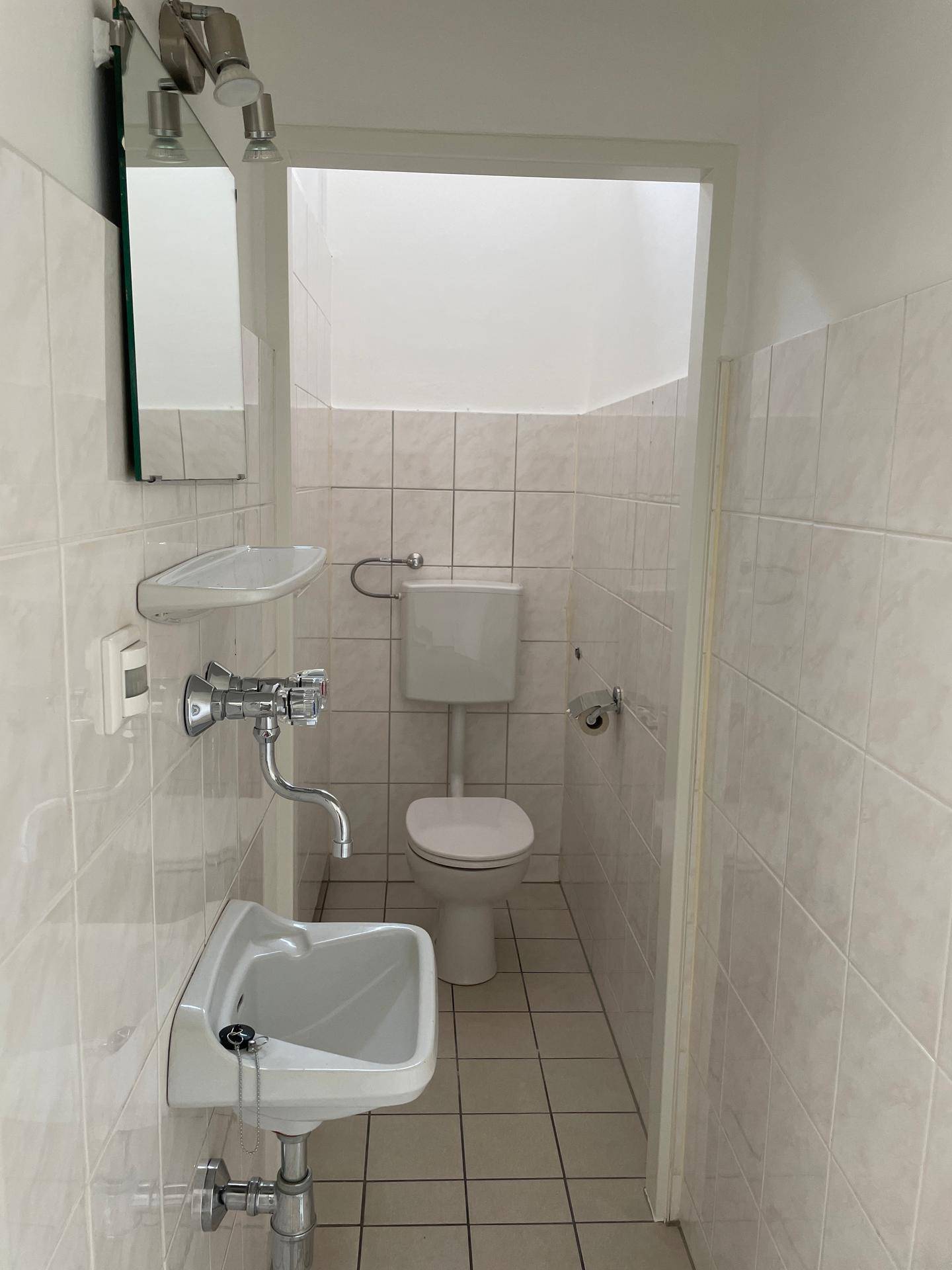 Schmaler Toilettenraum mit weiß gefliesten Wänden und Boden, ausgestattet mit WC und Waschbecken.