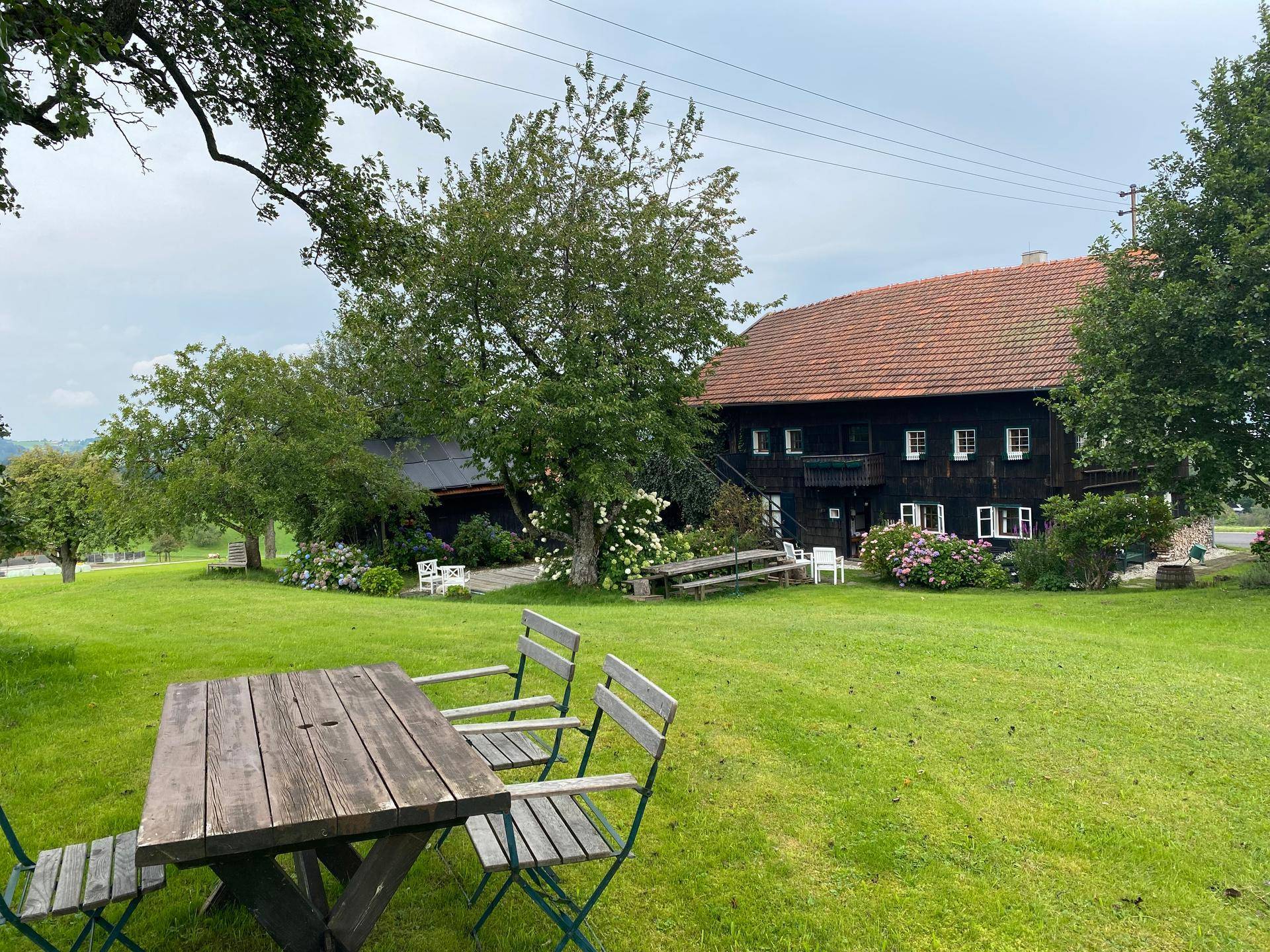 Großer Garten mit Holztisch und Stühlen vor einem traditionellen Bauernhaus mit rotem Ziegeldach.