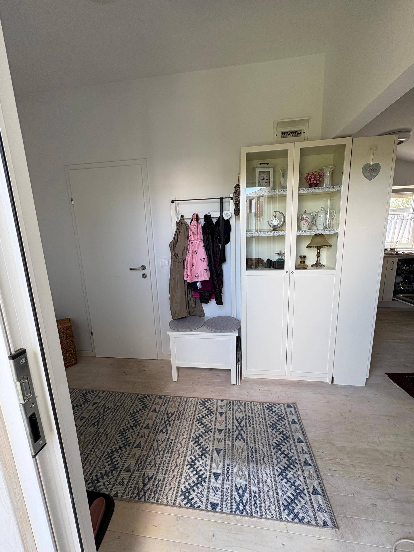 Heller Eingangsbereich mit Garderobe, Sitzbank und einem Vitrinenschrank auf hellem Holzboden.