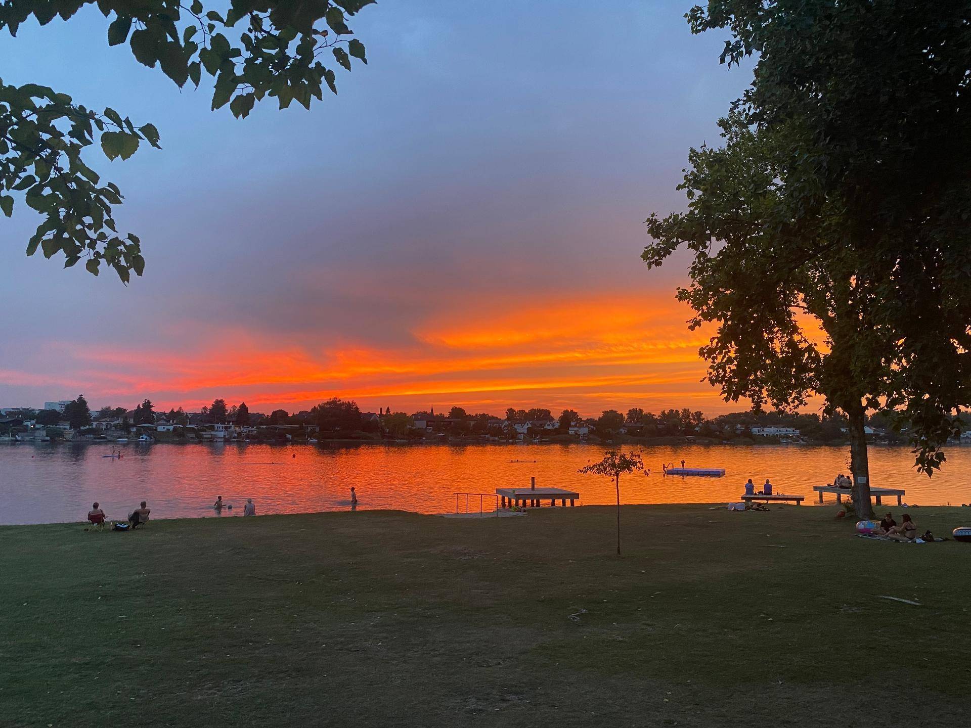 Spektakulärer Sonnenuntergang über einem See mit Menschen, die am Ufer entspannen und schwimmen.