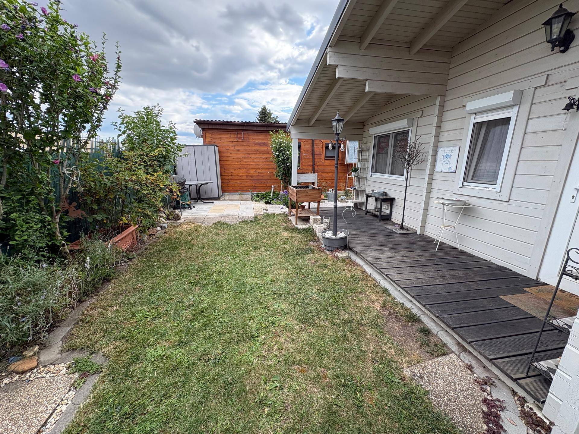 Seitlicher Gartenbereich mit Rasen, Holzterrasse und einem kleinen Gartenhaus im Hintergrund.