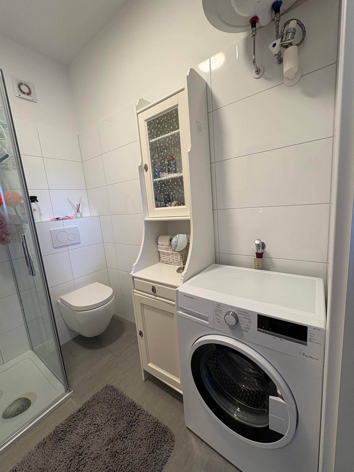 Modernes Badezimmer mit Dusche, WC und integrierter Waschmaschine auf Fliesenboden.