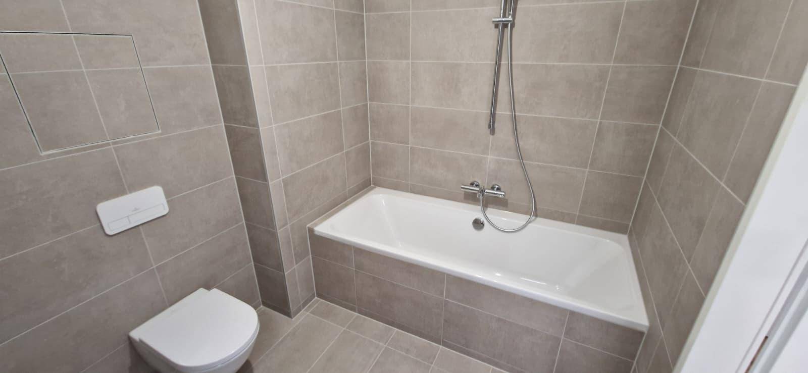 Zeitgemäßes Badezimmer mit Badewanne, Toilette und grauen Fliesen an Wänden und Boden.